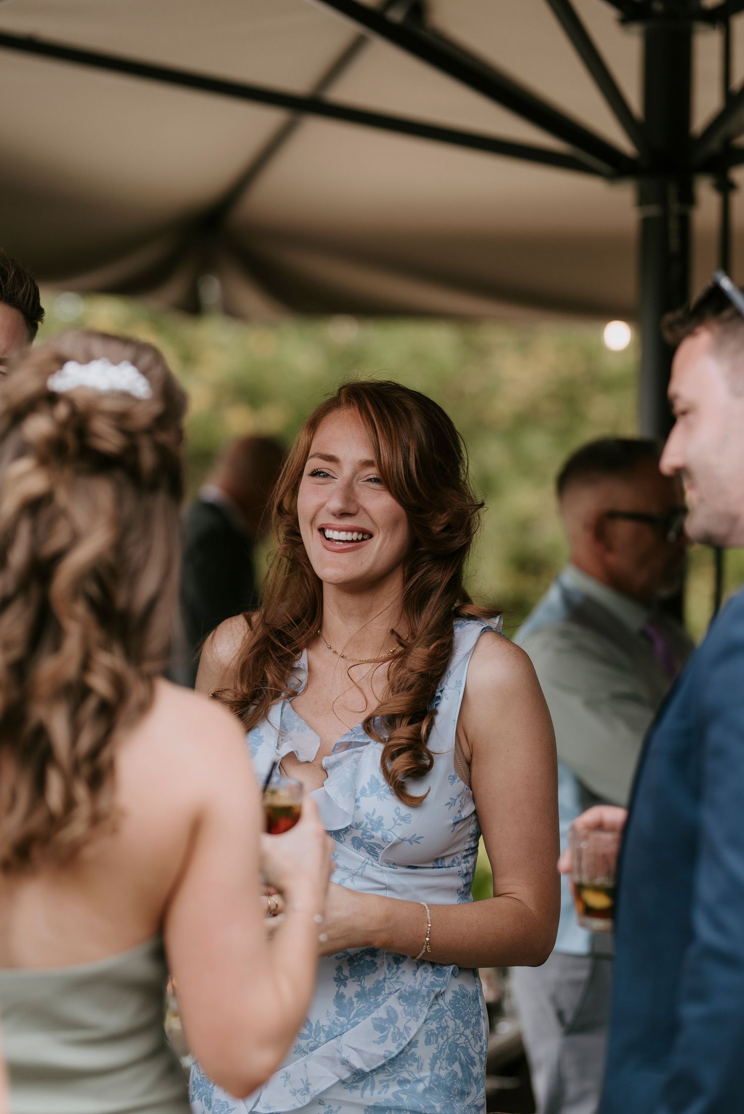 A group of wedding guests share laughter and conversation under a covered outdoor venue during a celebration.