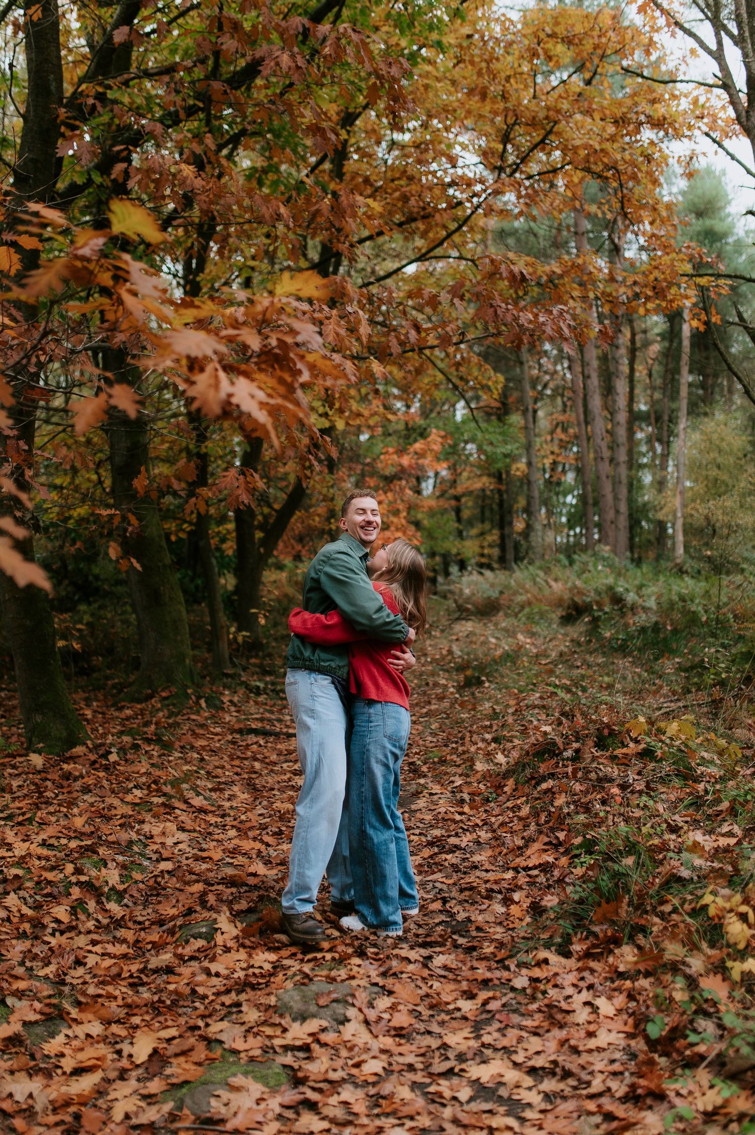A couple embraces in a forest covered in fallen autumn leaves during a romantic outdoor photoshoot.