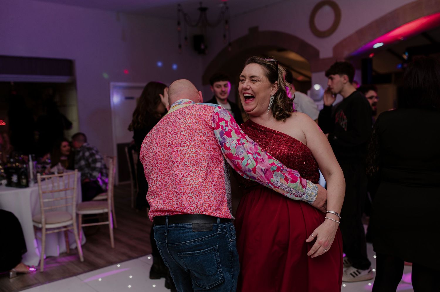 People dancing and celebrating at a wedding reception with colorful lighting and decorations.