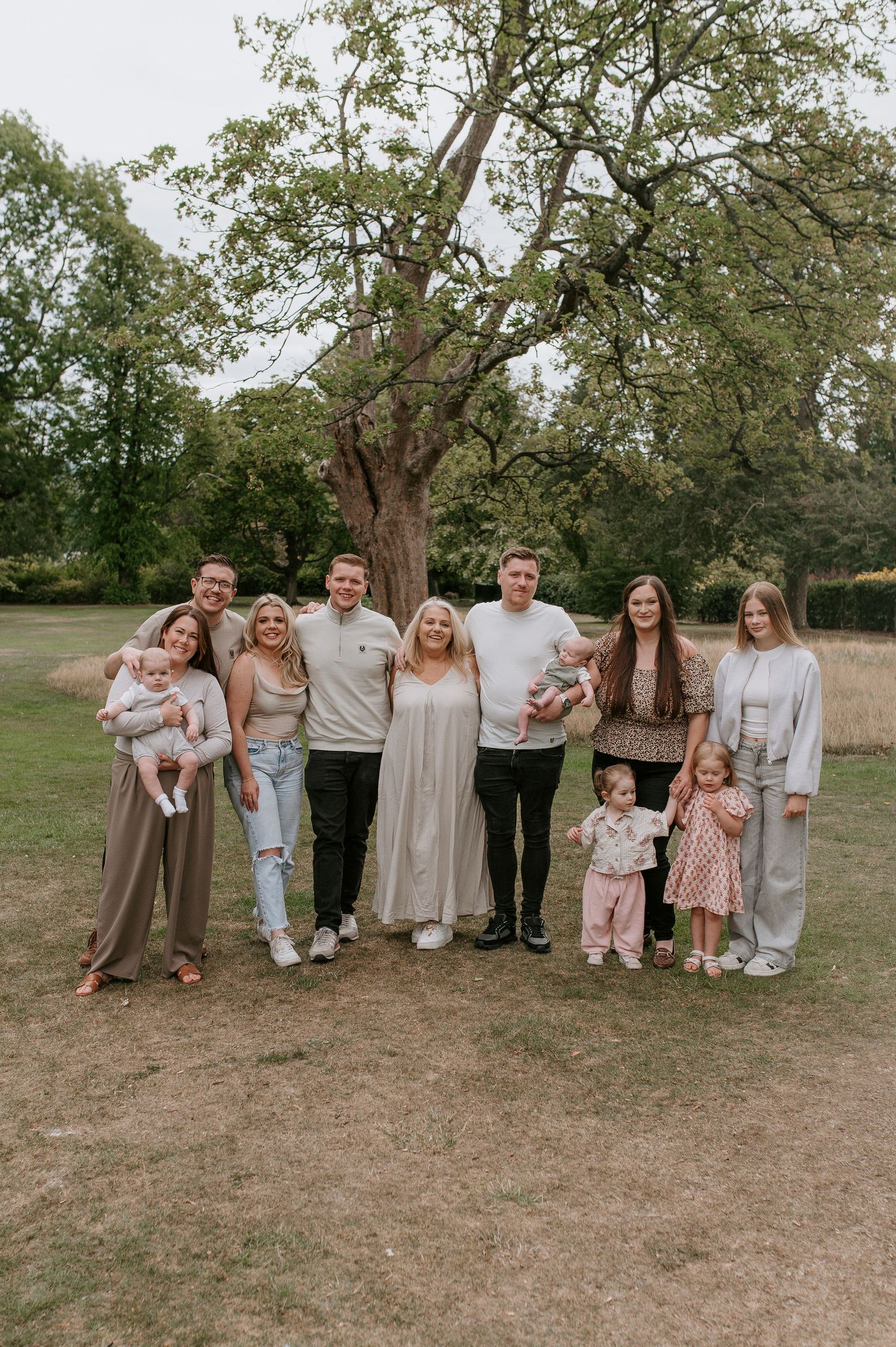 A group wearing coordinated neutral-toned outfits poses together on a grassy field during an outdoor photo session.