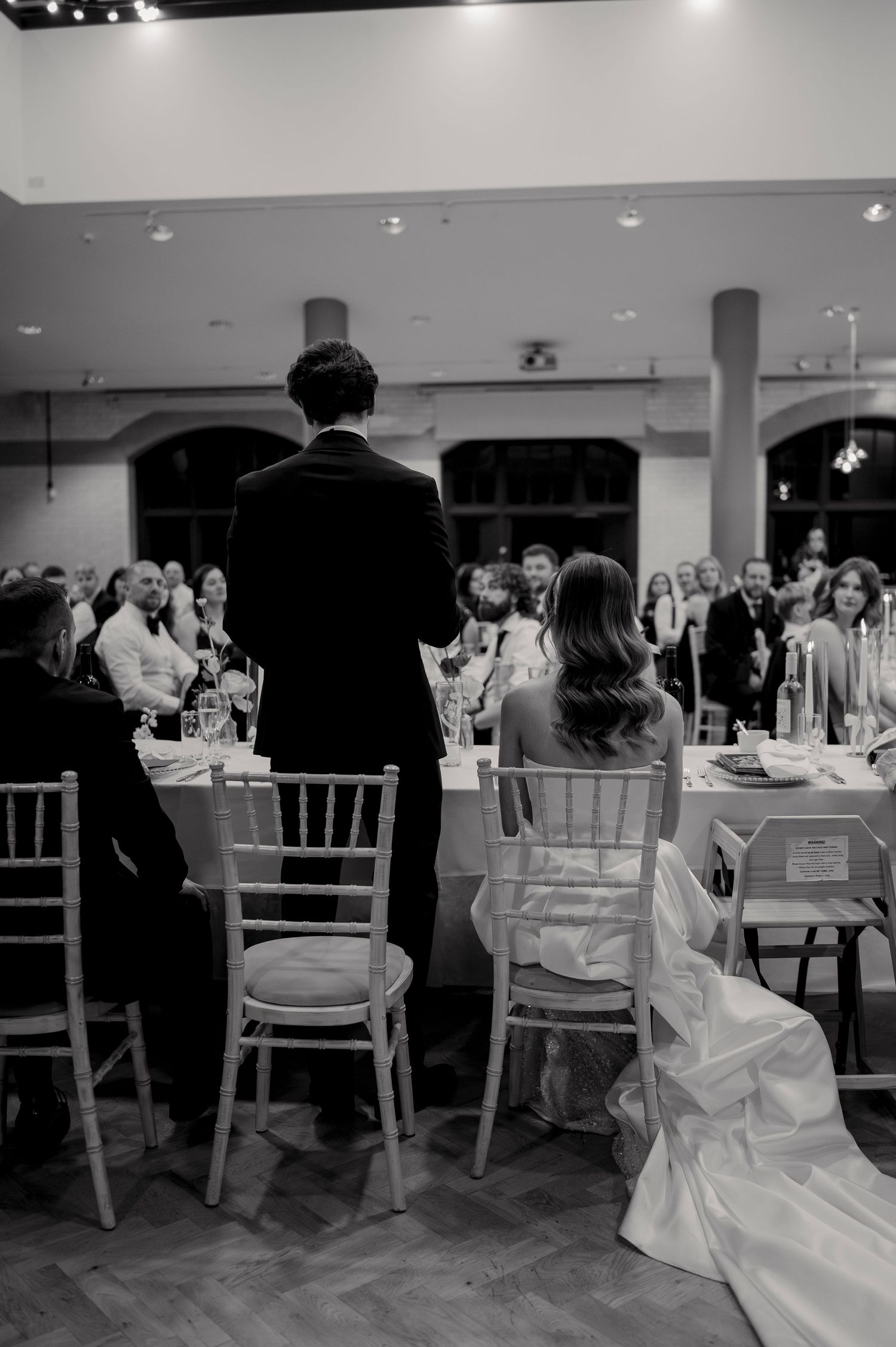 Black and white photo of a wedding reception with guests seated at tables while a speaker addresses the crowd.