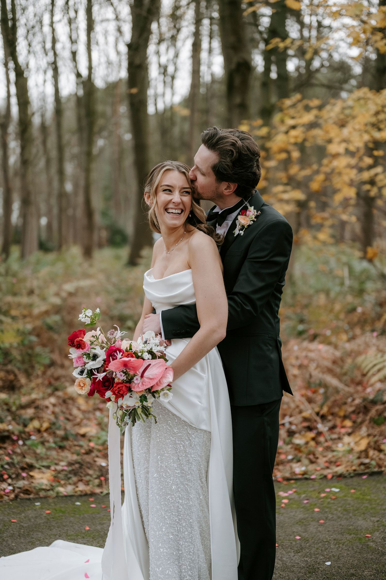 Newlyweds embrace in autumn forest setting with bride holding vibrant pink and red bouquet.
