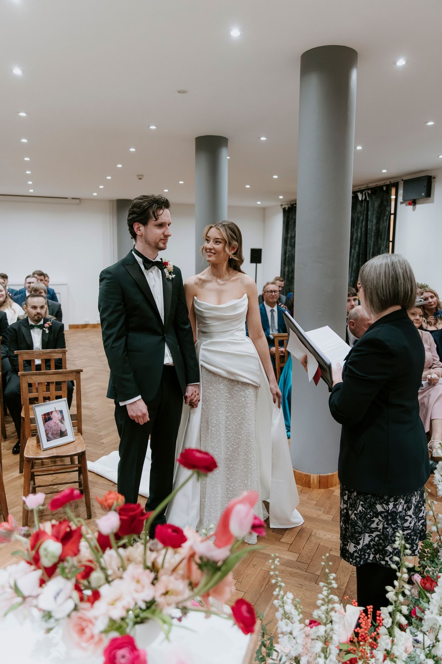 Wedding reception space with pink floral arrangements and guests mingling around white columns and wooden furniture.