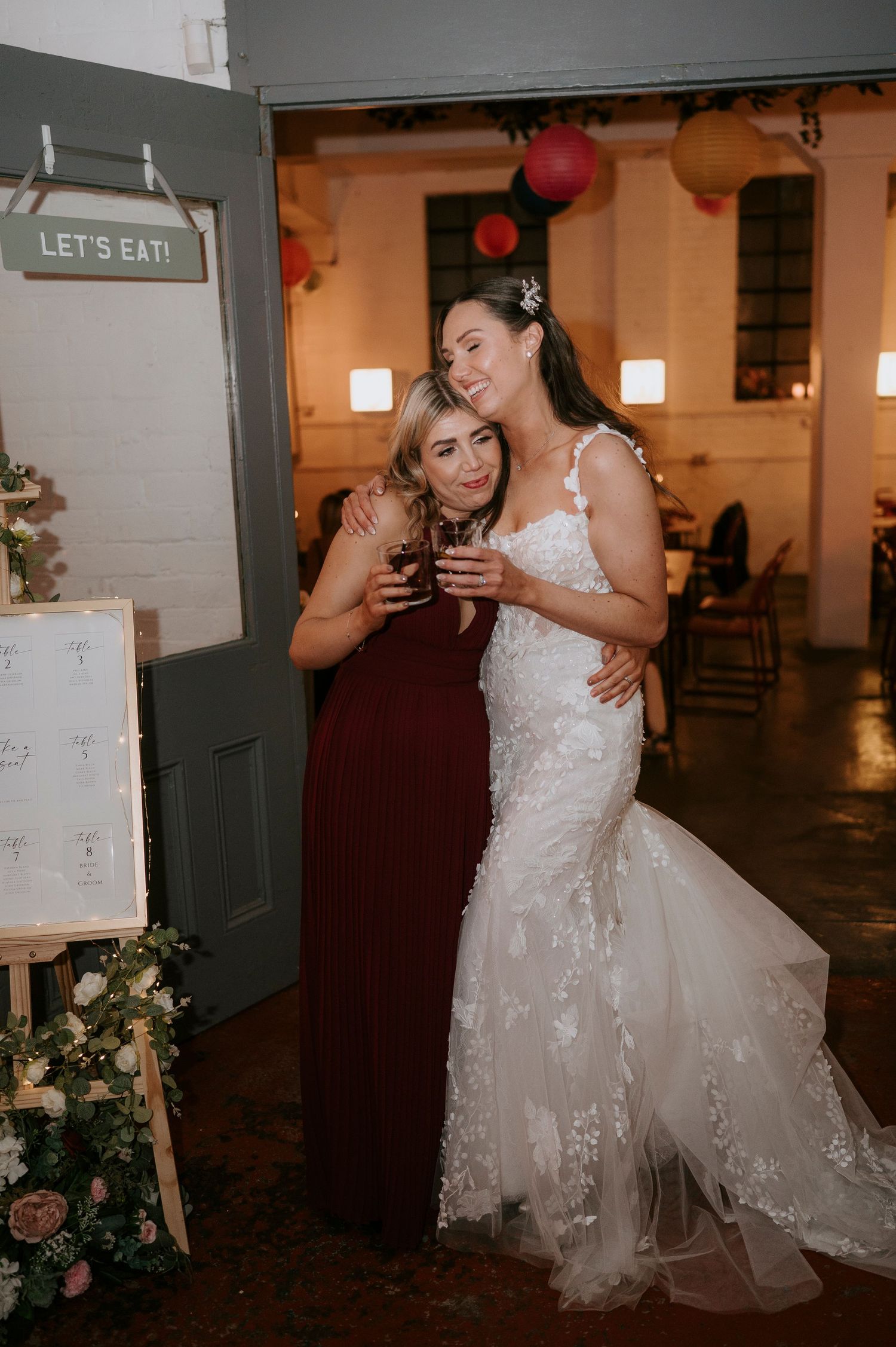 Bride in lace gown poses with bridesmaid in burgundy dress near wedding seating chart.