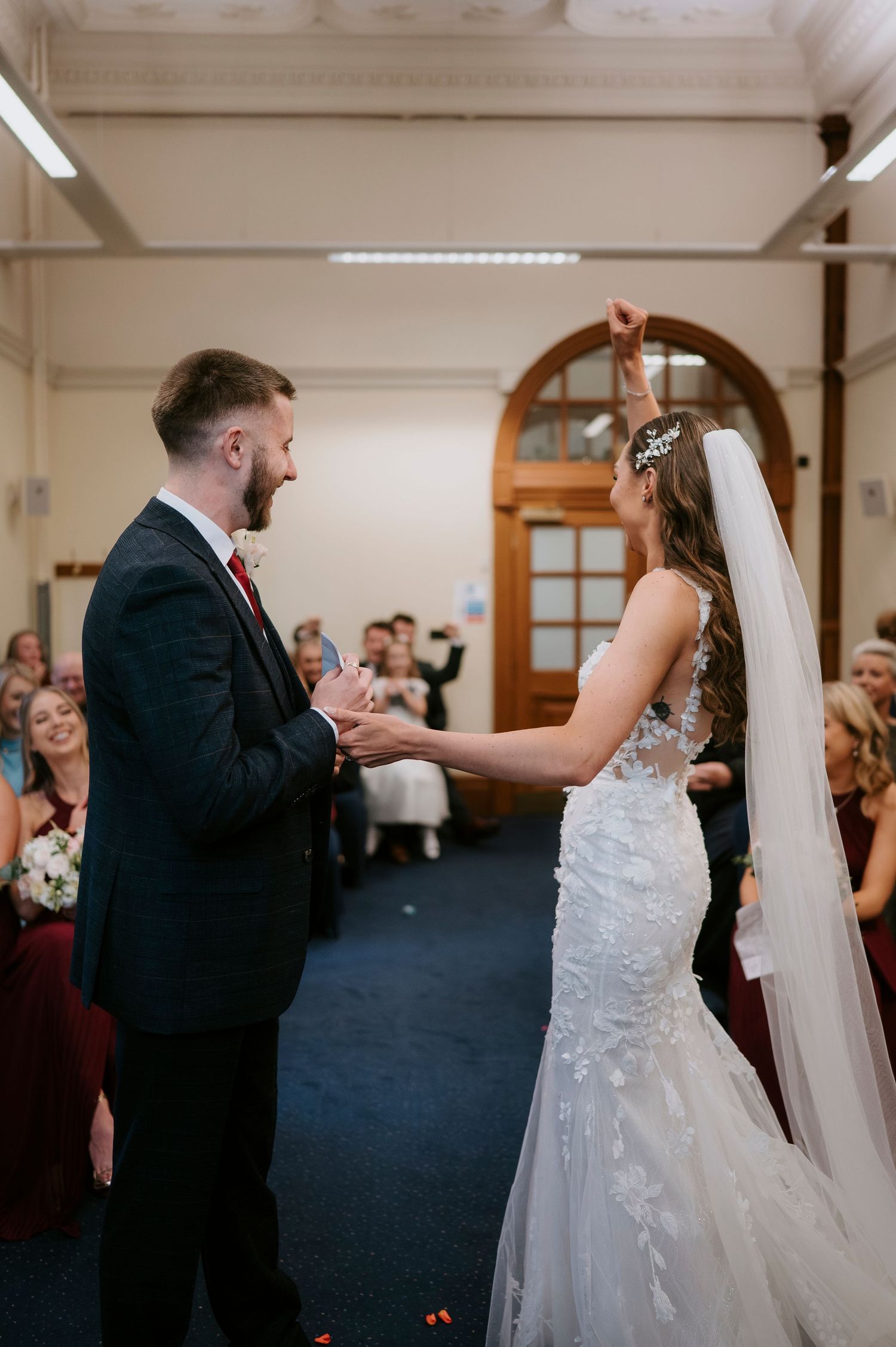 Bride and groom holding hands during wedding ceremony in elegant venue with guests seated in background.