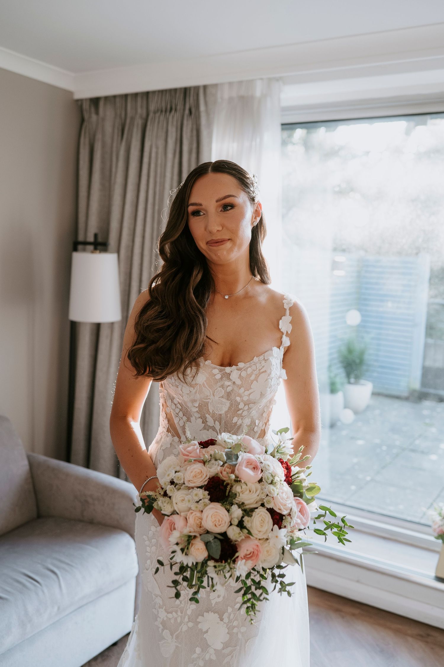 Bride in lace wedding gown holding pink and white bouquet standing by window in bridal suite.