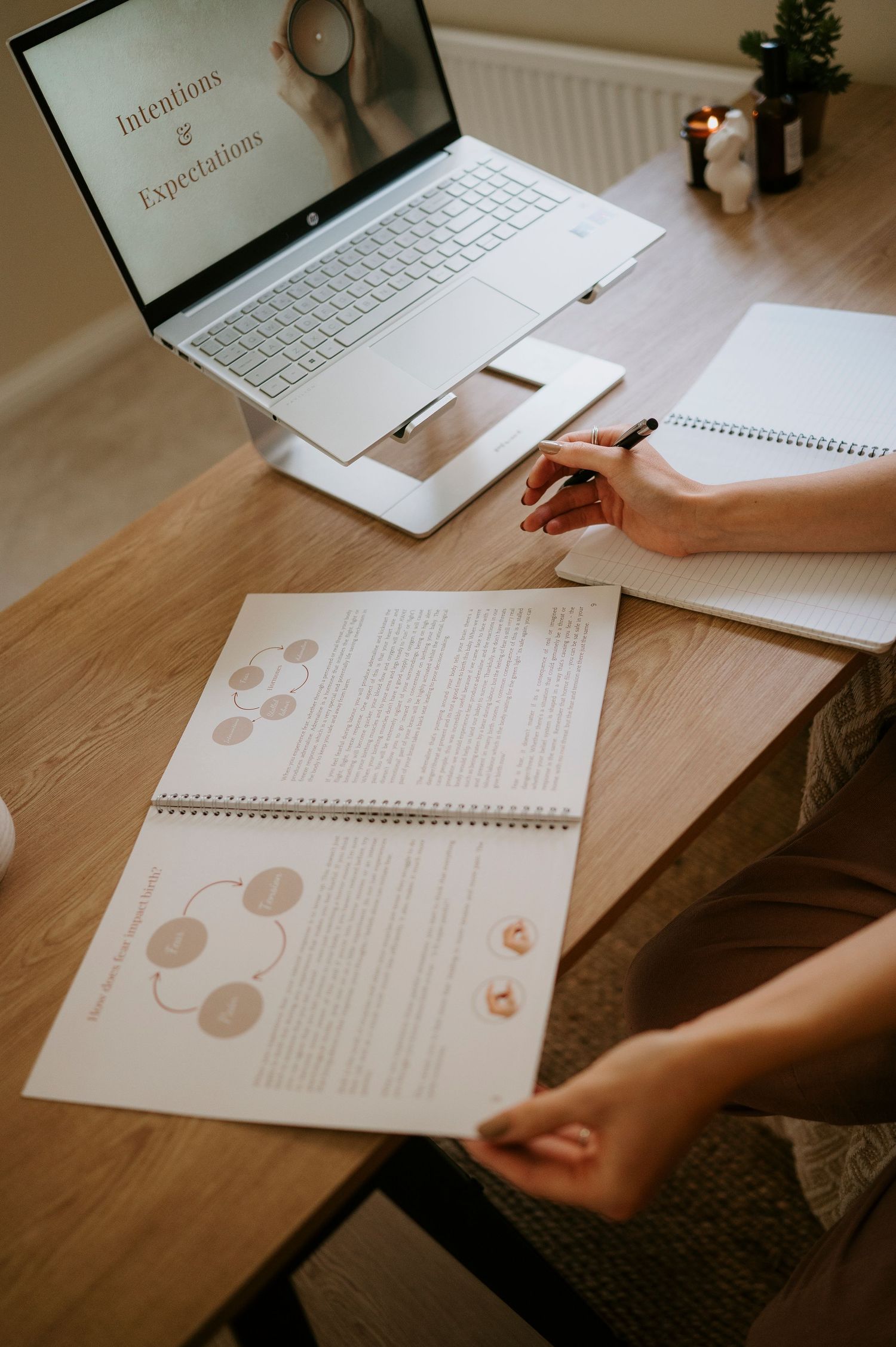 A person writes in a notebook while working at a wooden desk with a laptop and planner visible in the workspace.