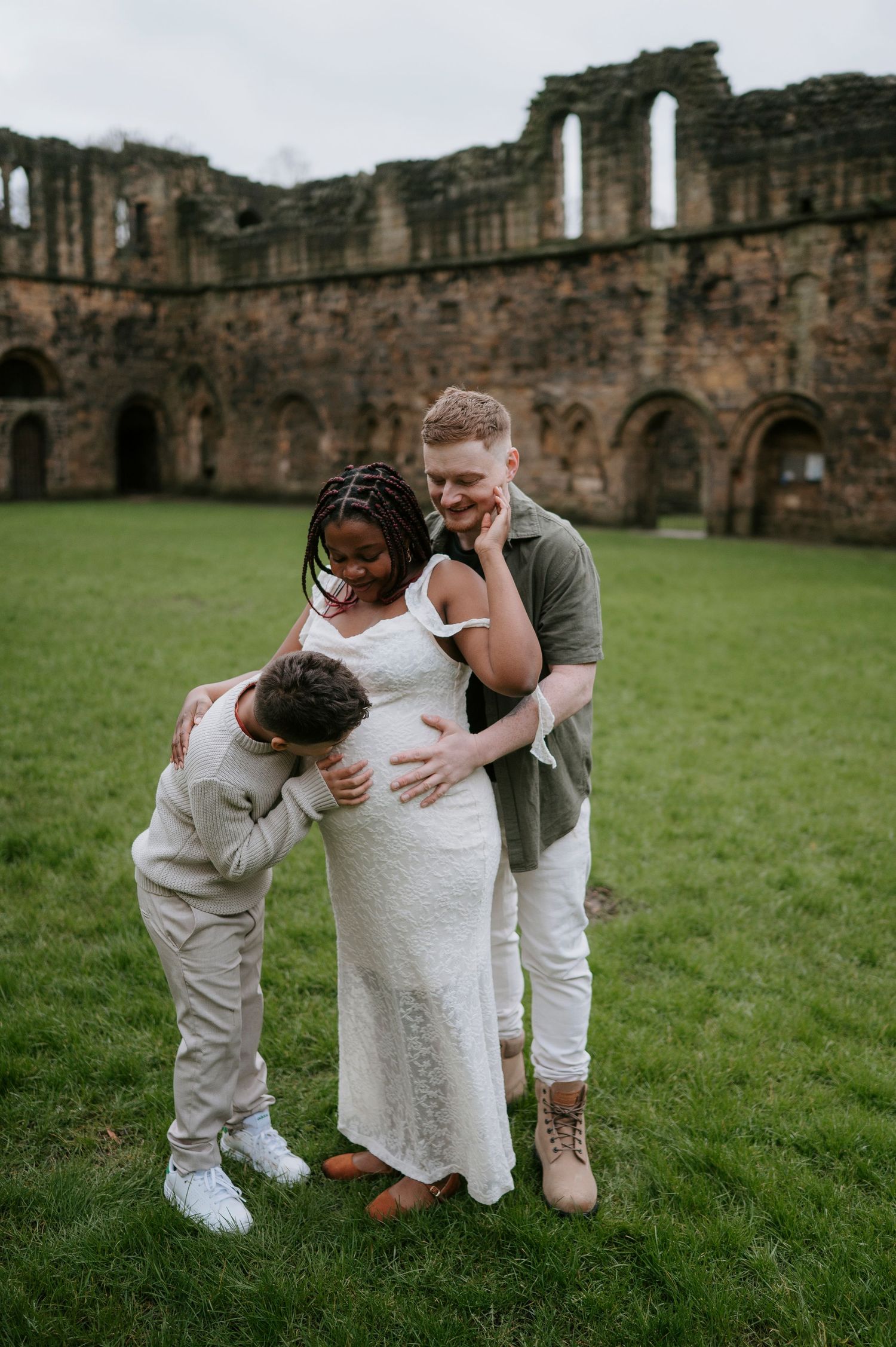 Three heartwarming photos show a touching family moment on green grass in front of historic castle ruins.