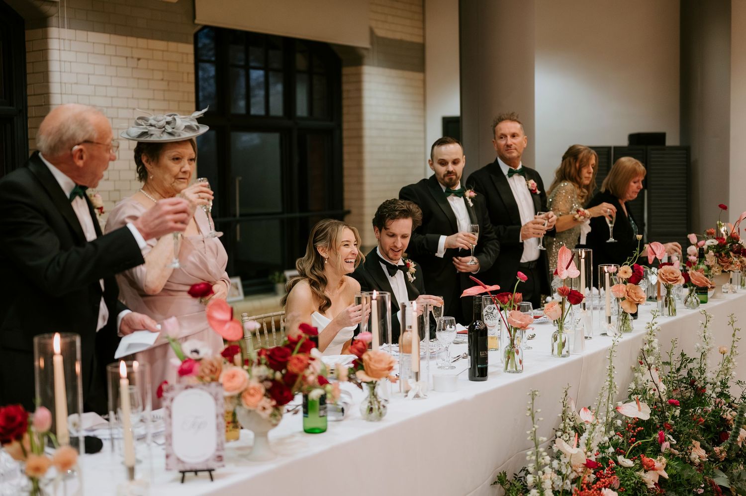 Wedding guests seated at a long reception table decorated with floral arrangements and drinks during a celebration.