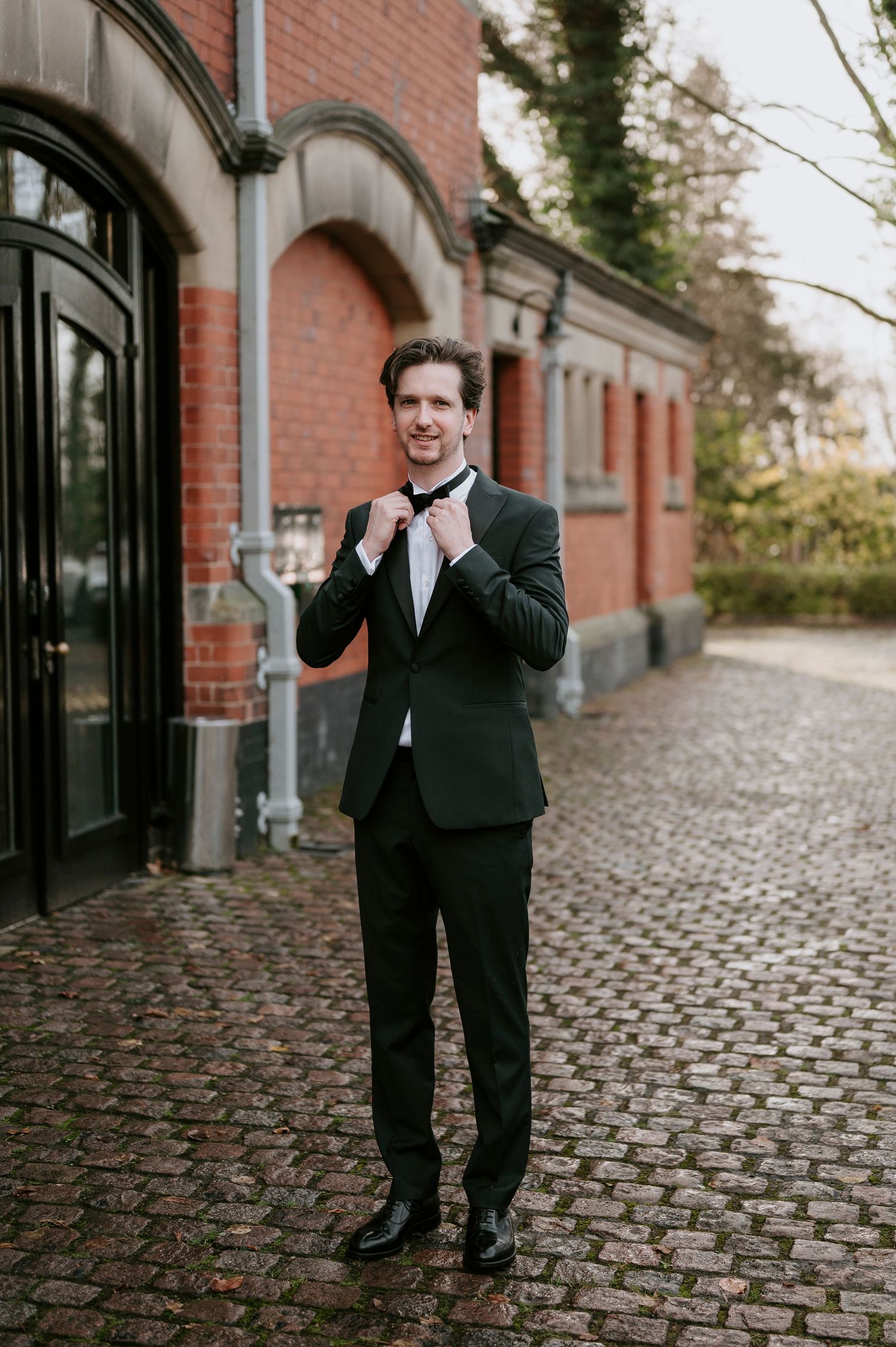 A person in a dark suit walks and poses playfully on a cobblestone path near a brick building.