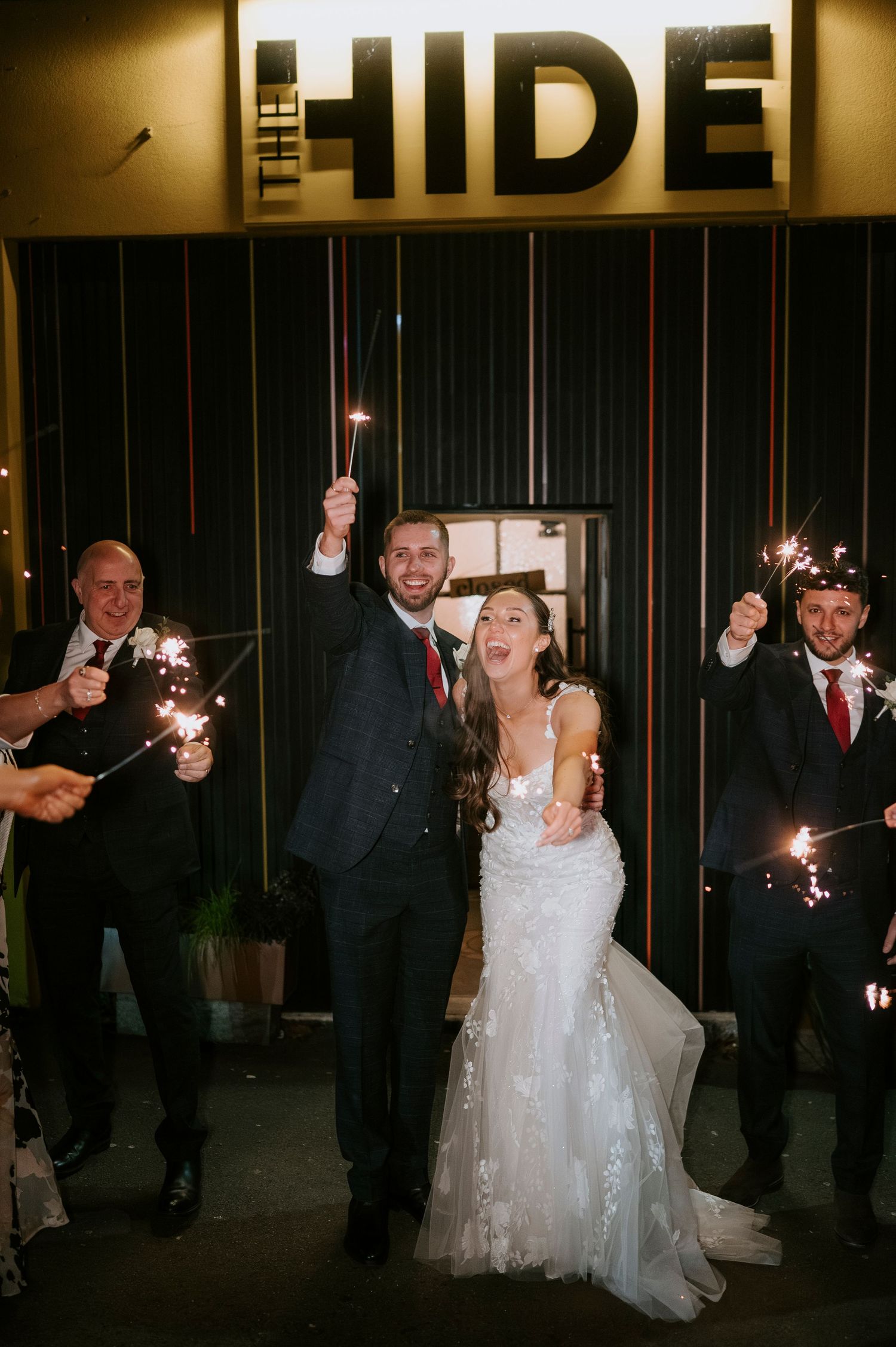 Bride and groom celebrate with sparklers outside The Hide venue surrounded by wedding guests.