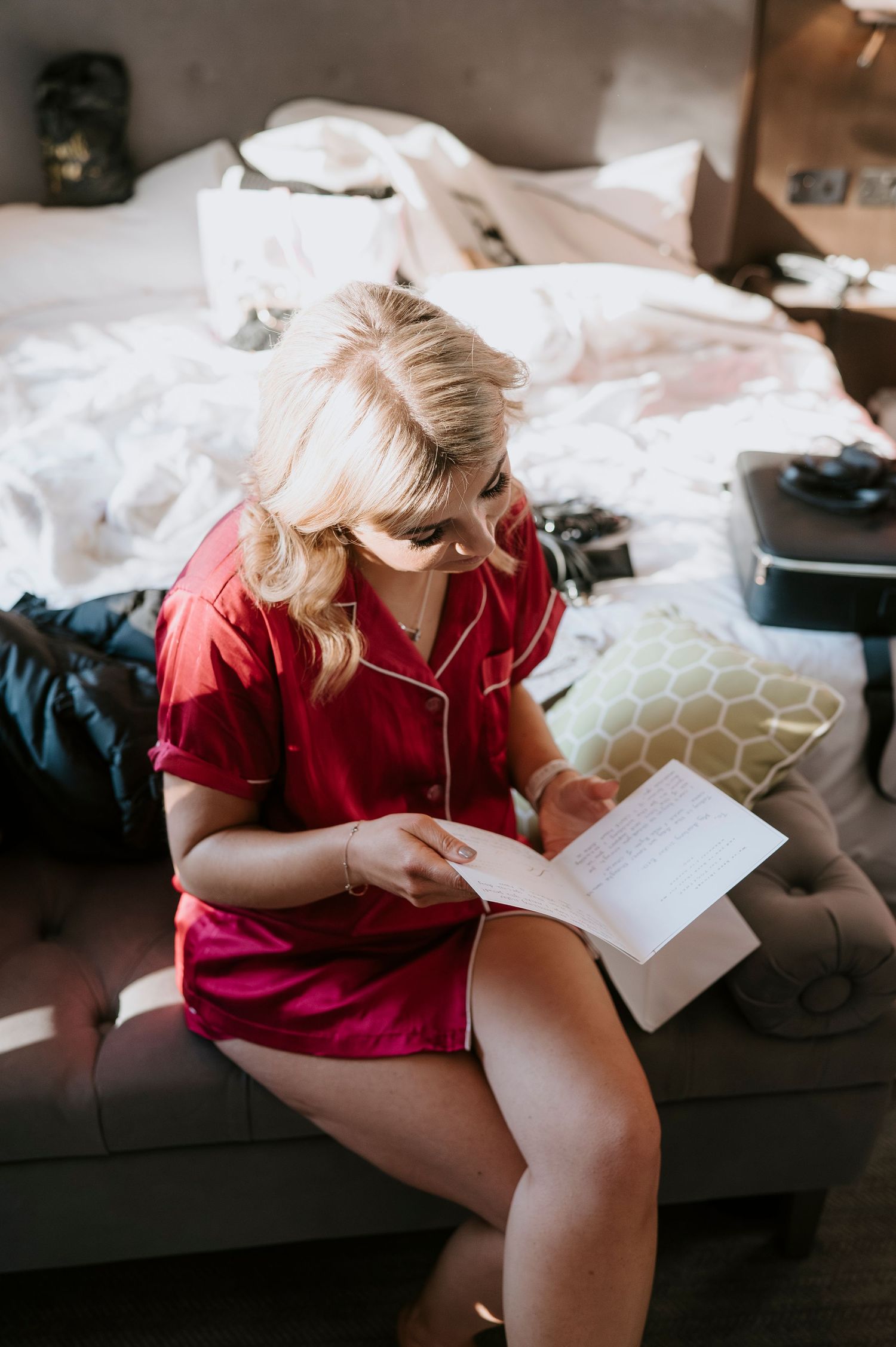 Blonde woman in red satin pajamas reading a handwritten letter while sitting on a couch in a cozy bedroom.