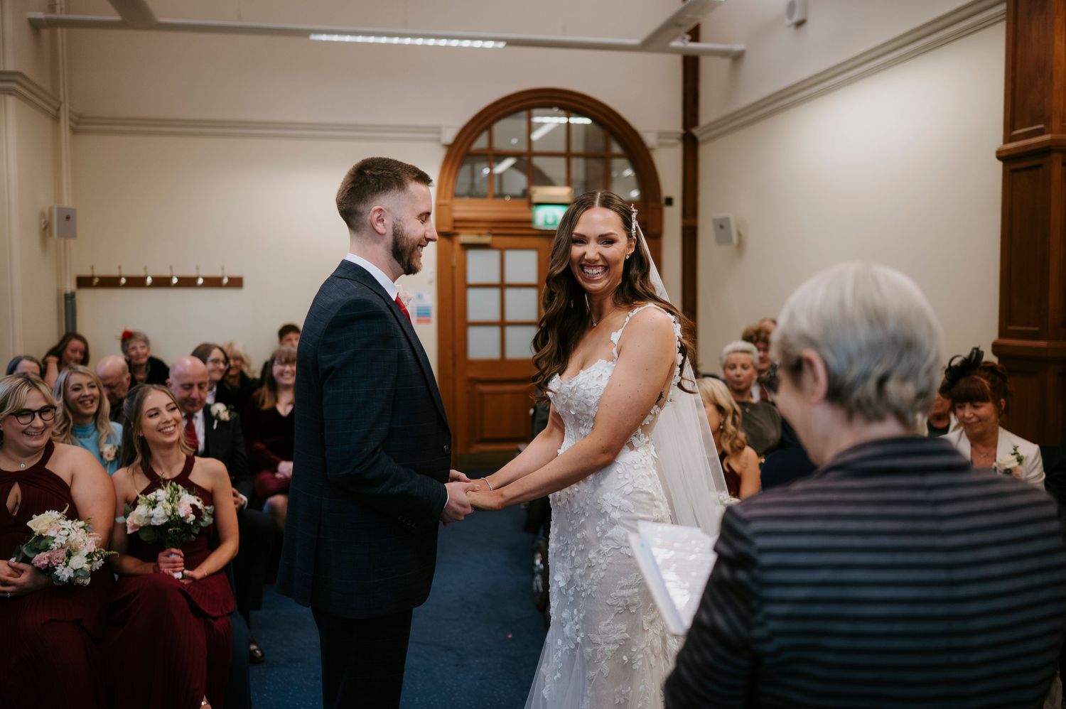 Bride and groom holding hands and smiling during wedding ceremony with guests seated in background.