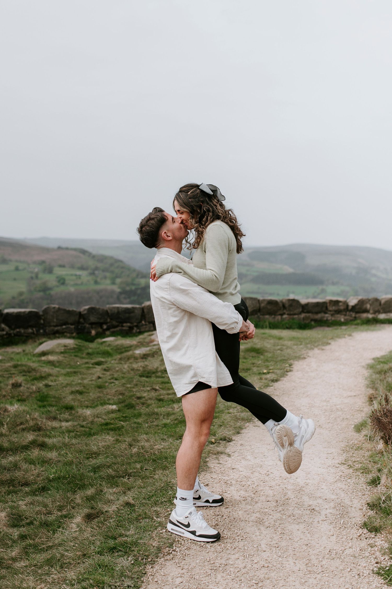 A couple shares a romantic embrace on a scenic hiking trail through rolling green hills on a cloudy day.