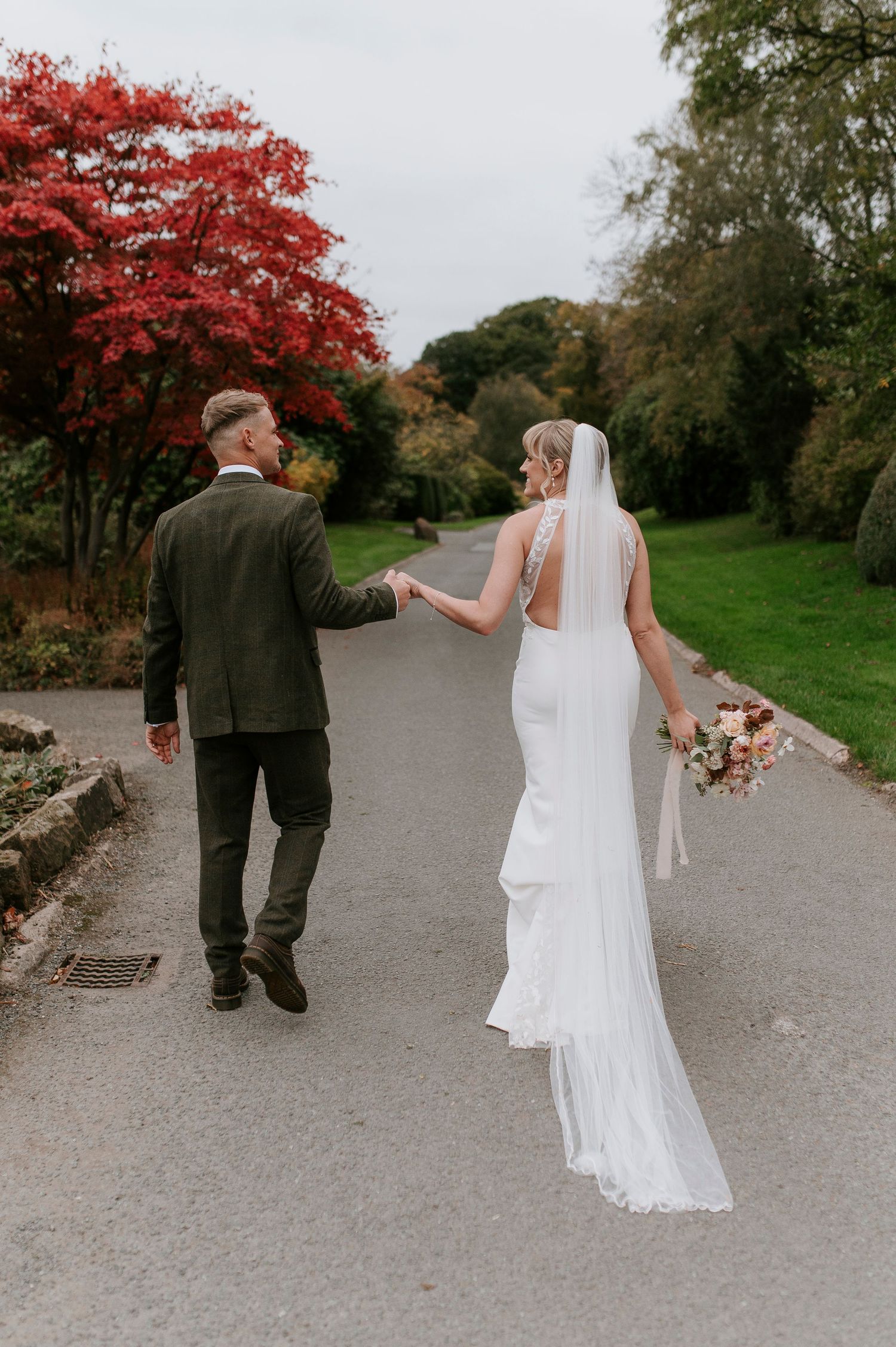 A couple in wedding attire walks along a gravel path surrounded by autumn trees with vibrant red foliage.