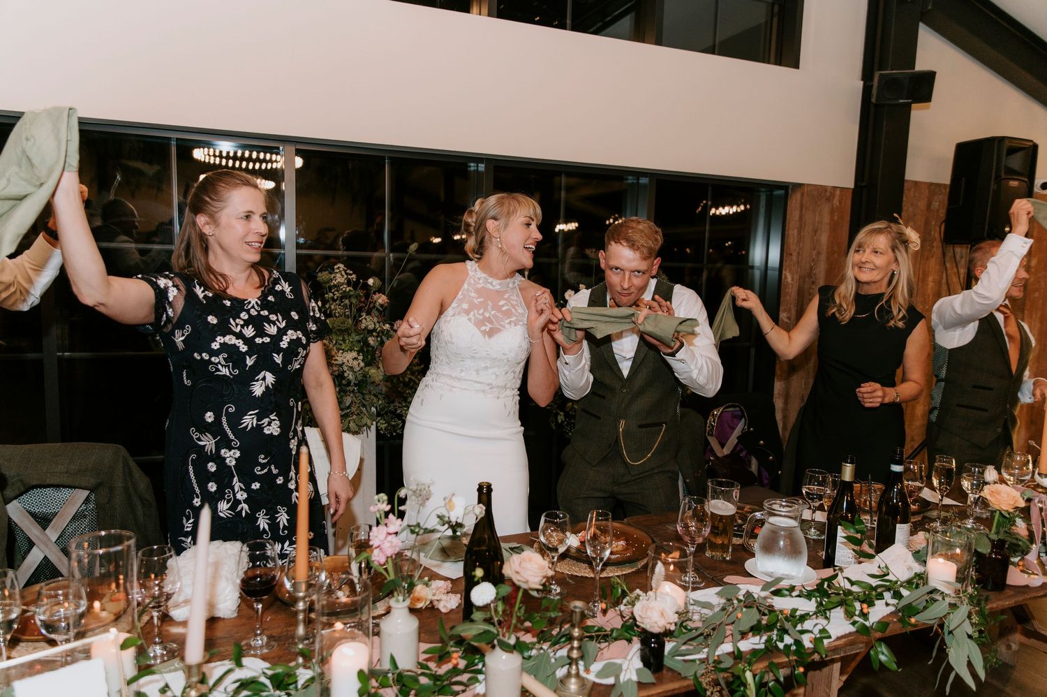 Wedding guests dance and celebrate together at a dimly lit indoor reception venue decorated with floral centerpieces.