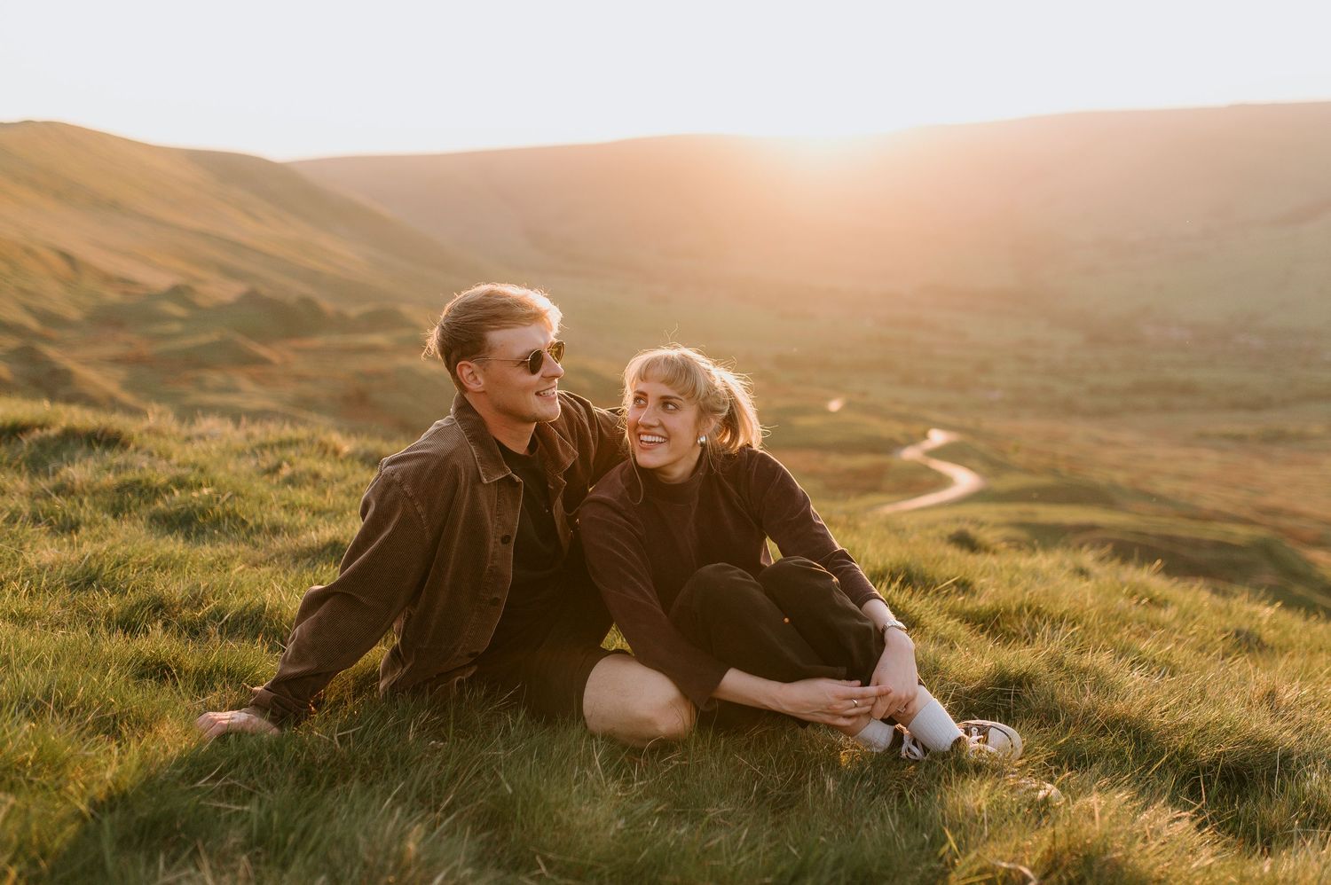 A couple sits together on a grassy hillside during golden hour with rolling hills in the background.