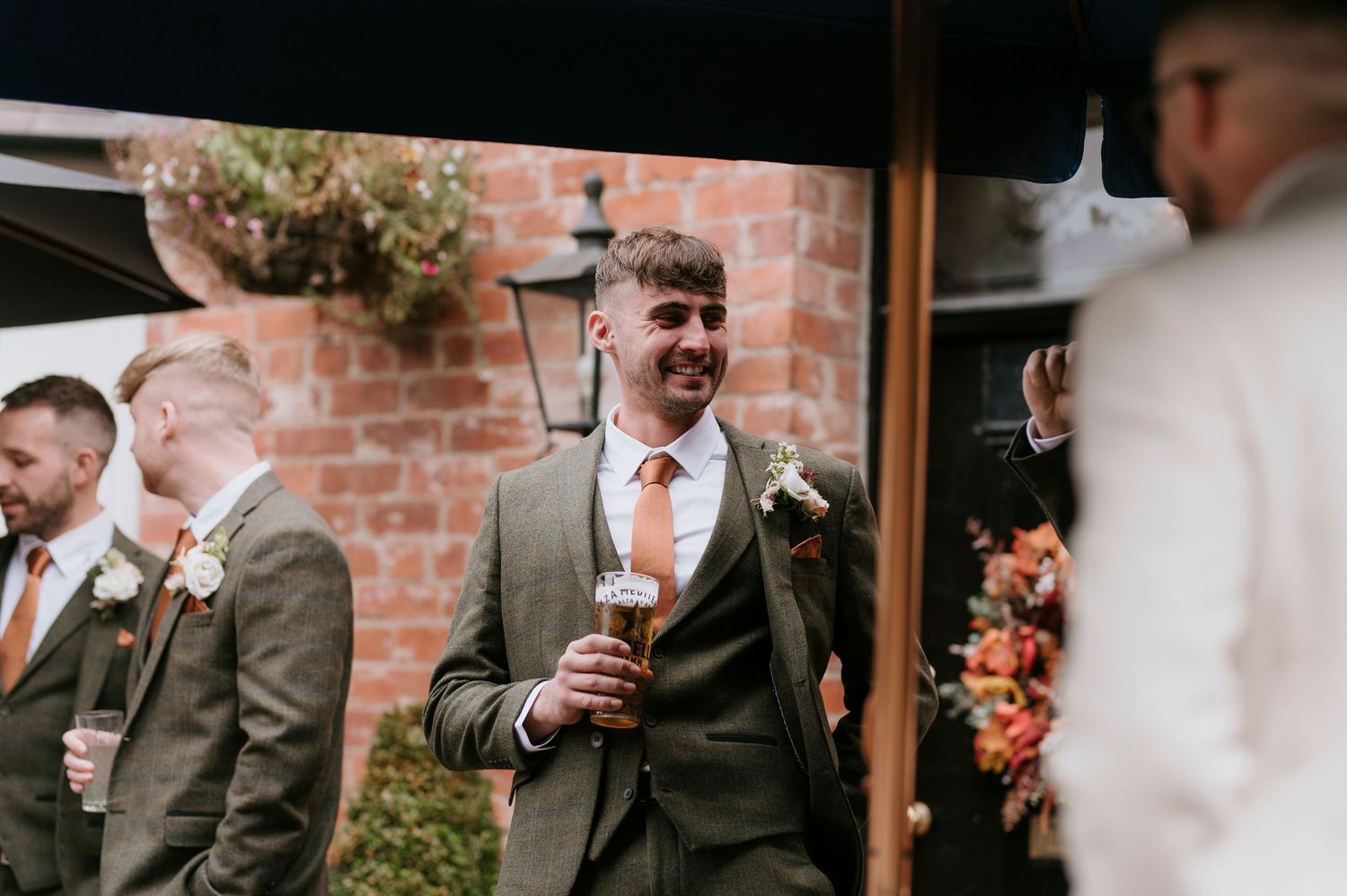 Wedding guests in tweed suits raise their glasses in a toast against a rustic brick wall backdrop.