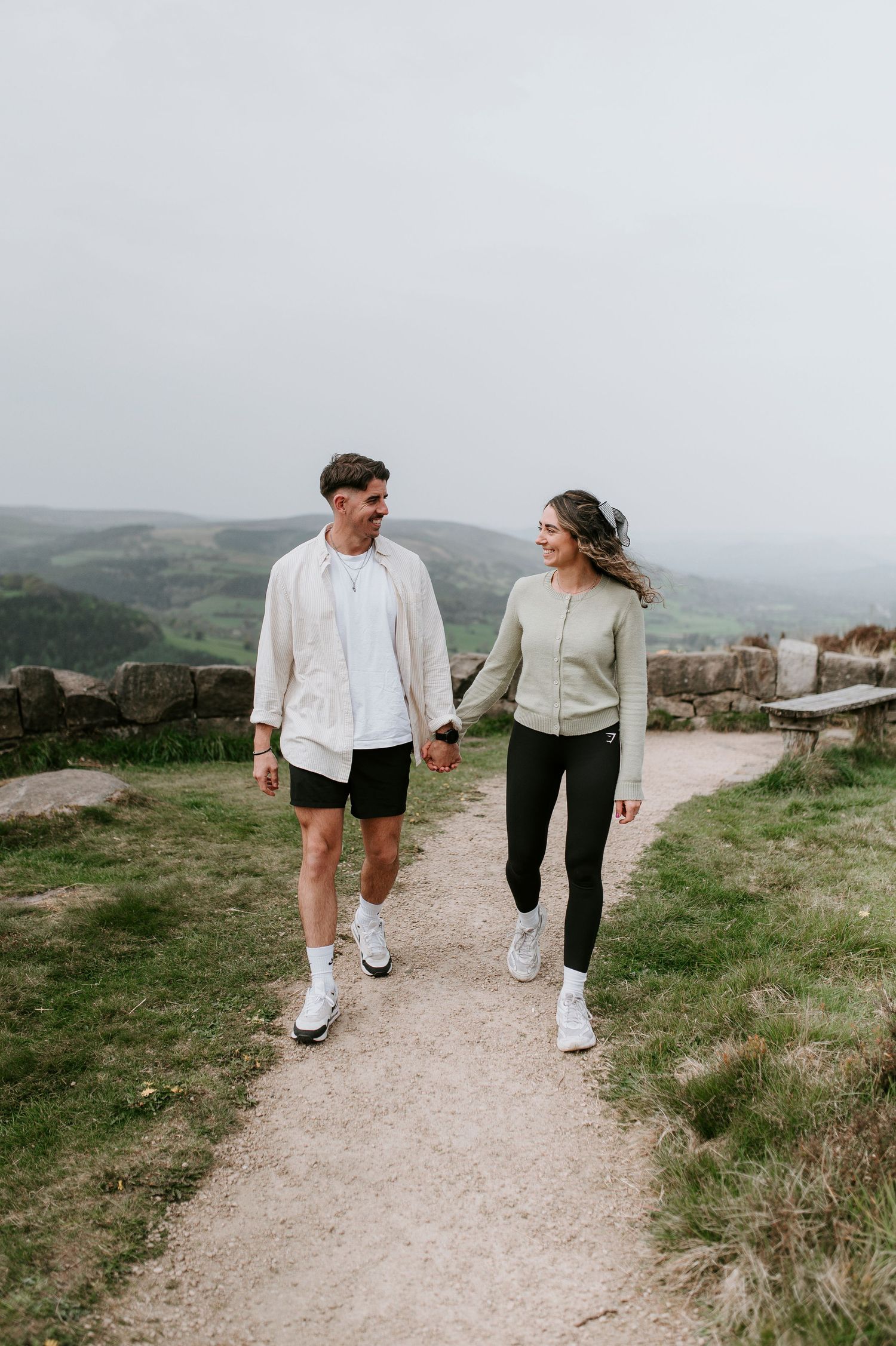 A couple walks together on a scenic coastal path wearing matching white sneakers and casual outfits.