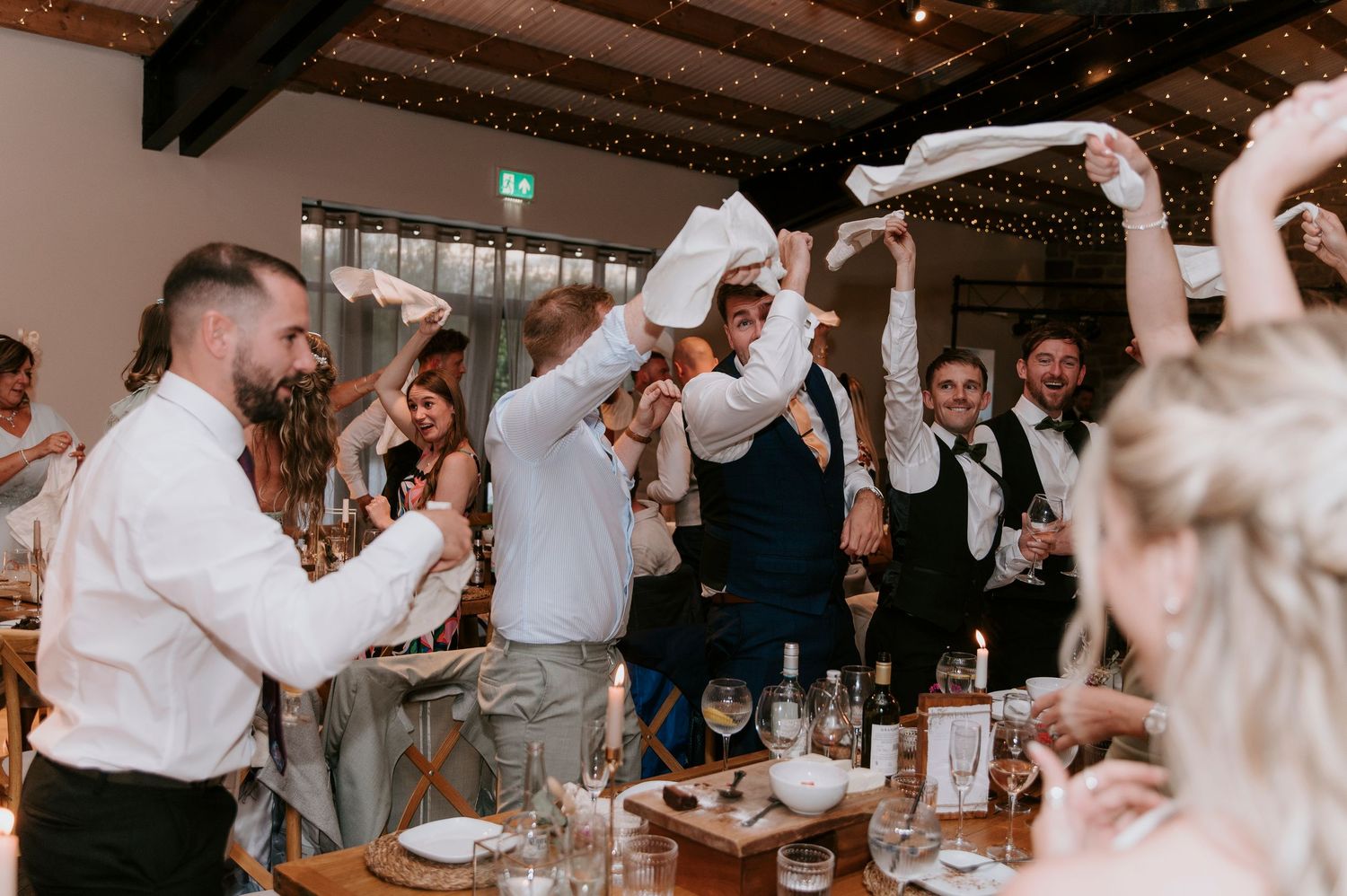 Wedding guests raise their arms and dance together during a lively reception celebration at an indoor venue.