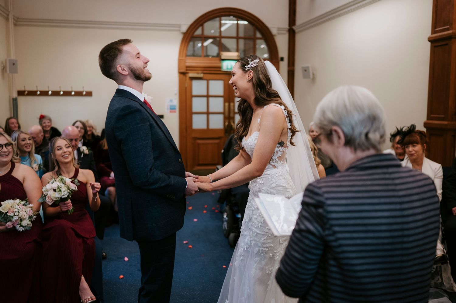 Bride and groom holding hands and smiling during indoor wedding ceremony with guests seated in background.