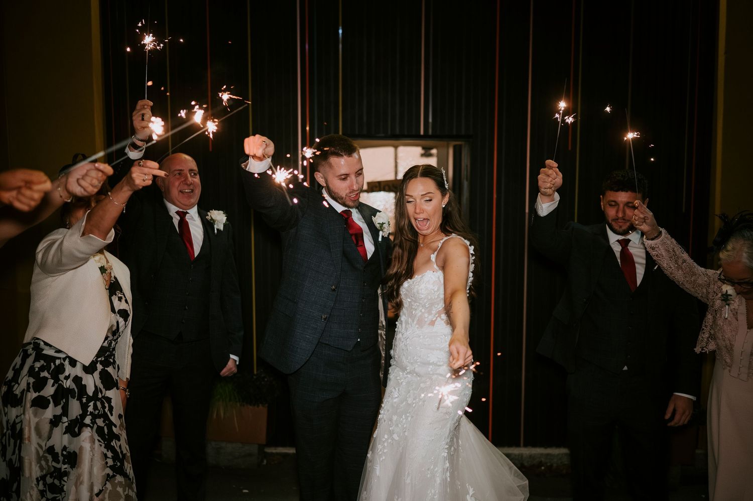 Newlyweds exit wedding venue through sparkler tunnel with family and guests celebrating.
