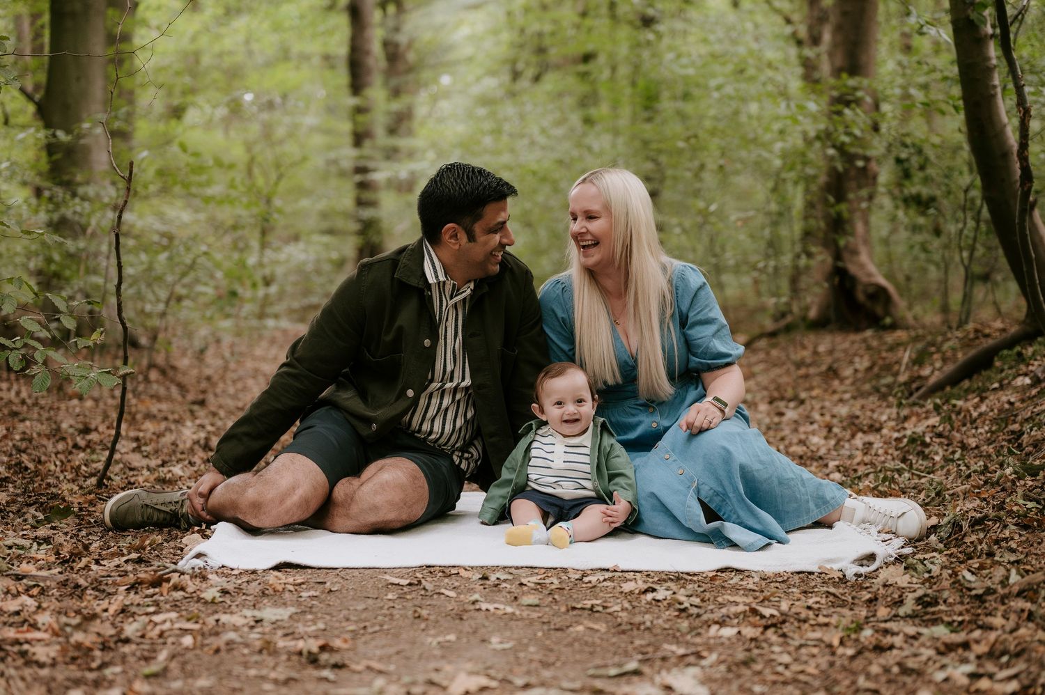 A couple and their child lie on a blanket in a serene forest setting during an outdoor family photoshoot.