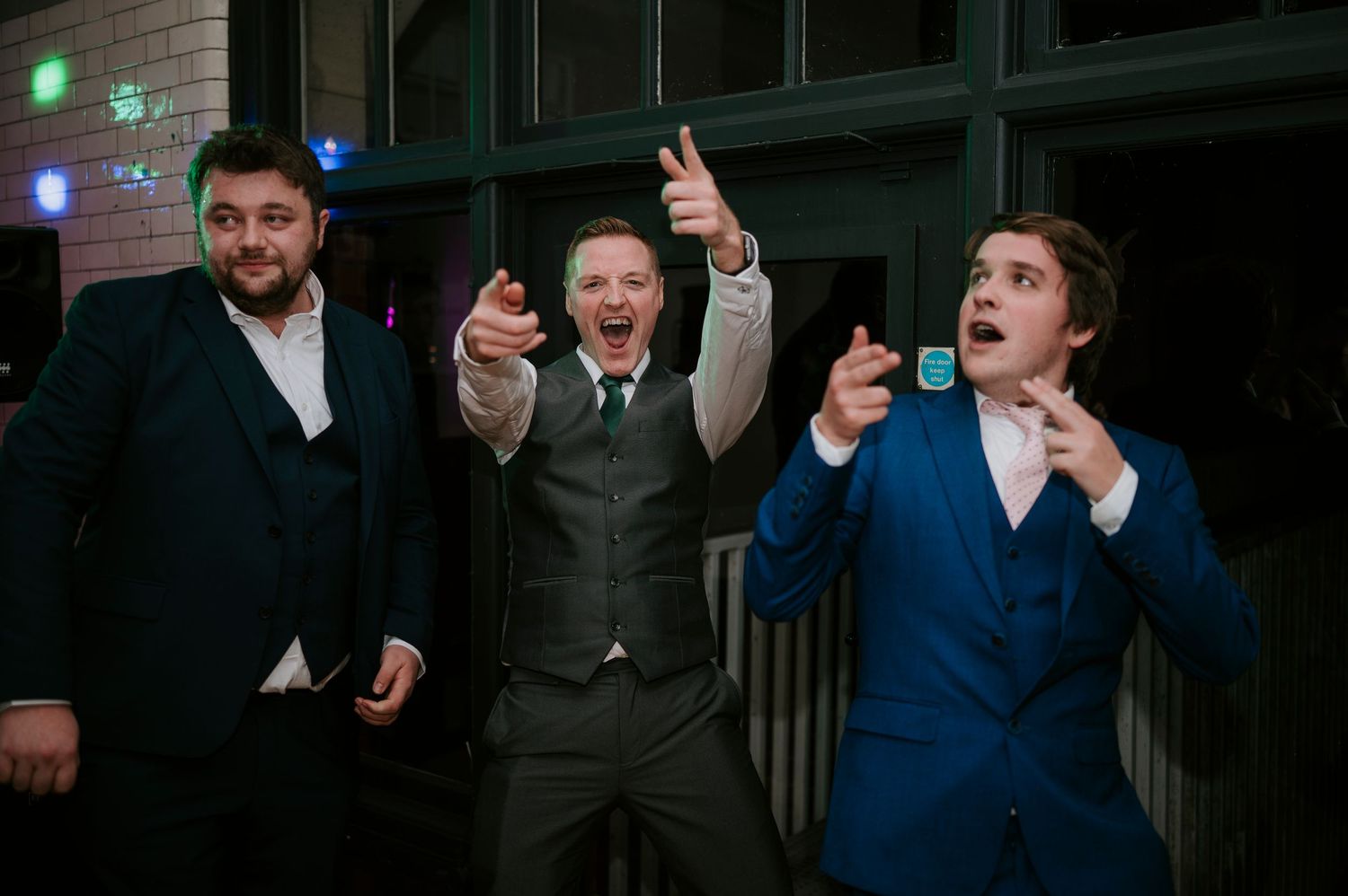 Wedding guests dance and celebrate with energetic expressions at an indoor reception.