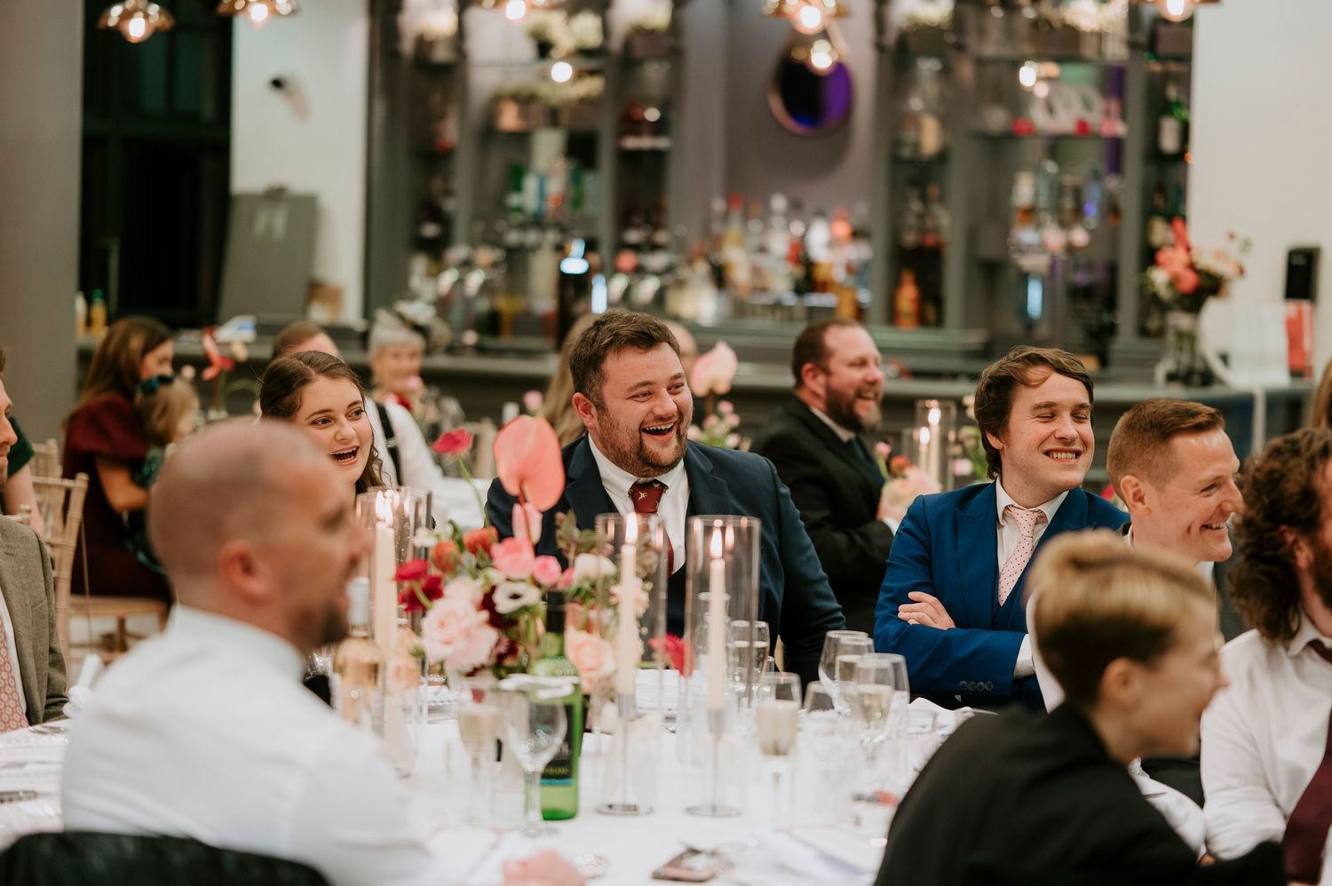 Wedding guests laugh and smile during a reception dinner at elegantly decorated tables with pink floral centerpieces.