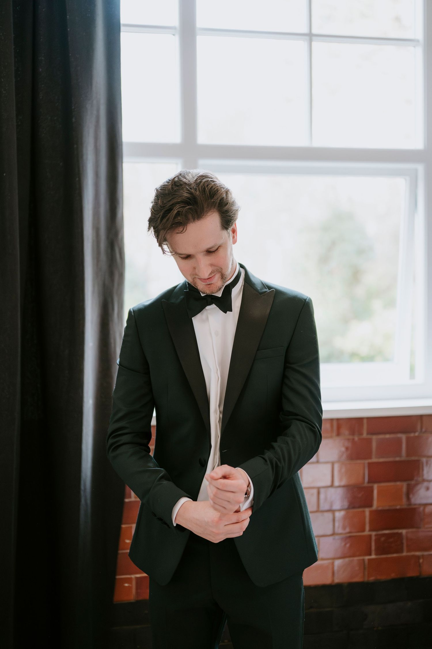 Groom in black tuxedo adjusts cuffs by window in brick-walled room.