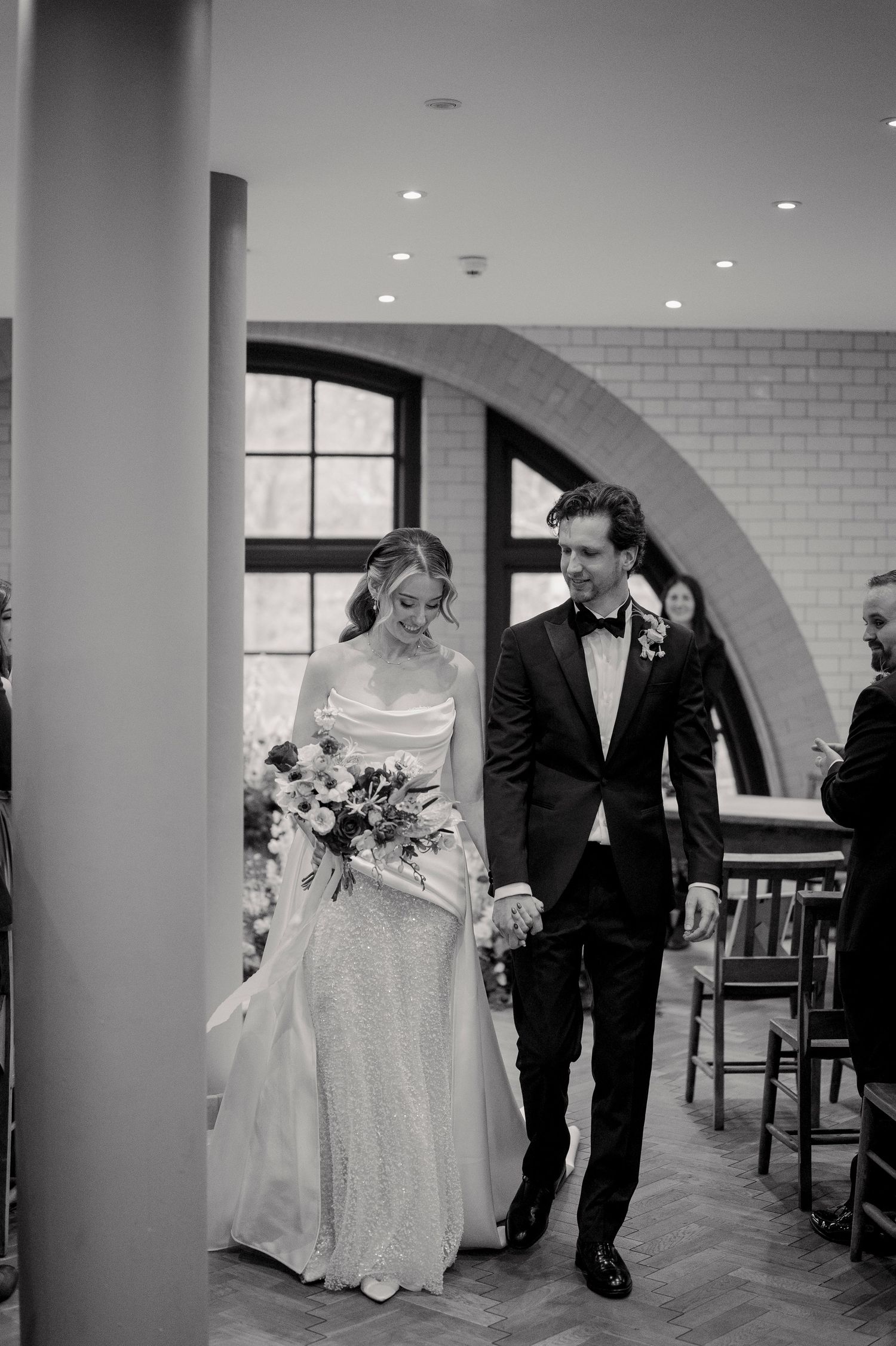 A wedding couple walks through an elegant arched hallway with white brick walls and recessed lighting.