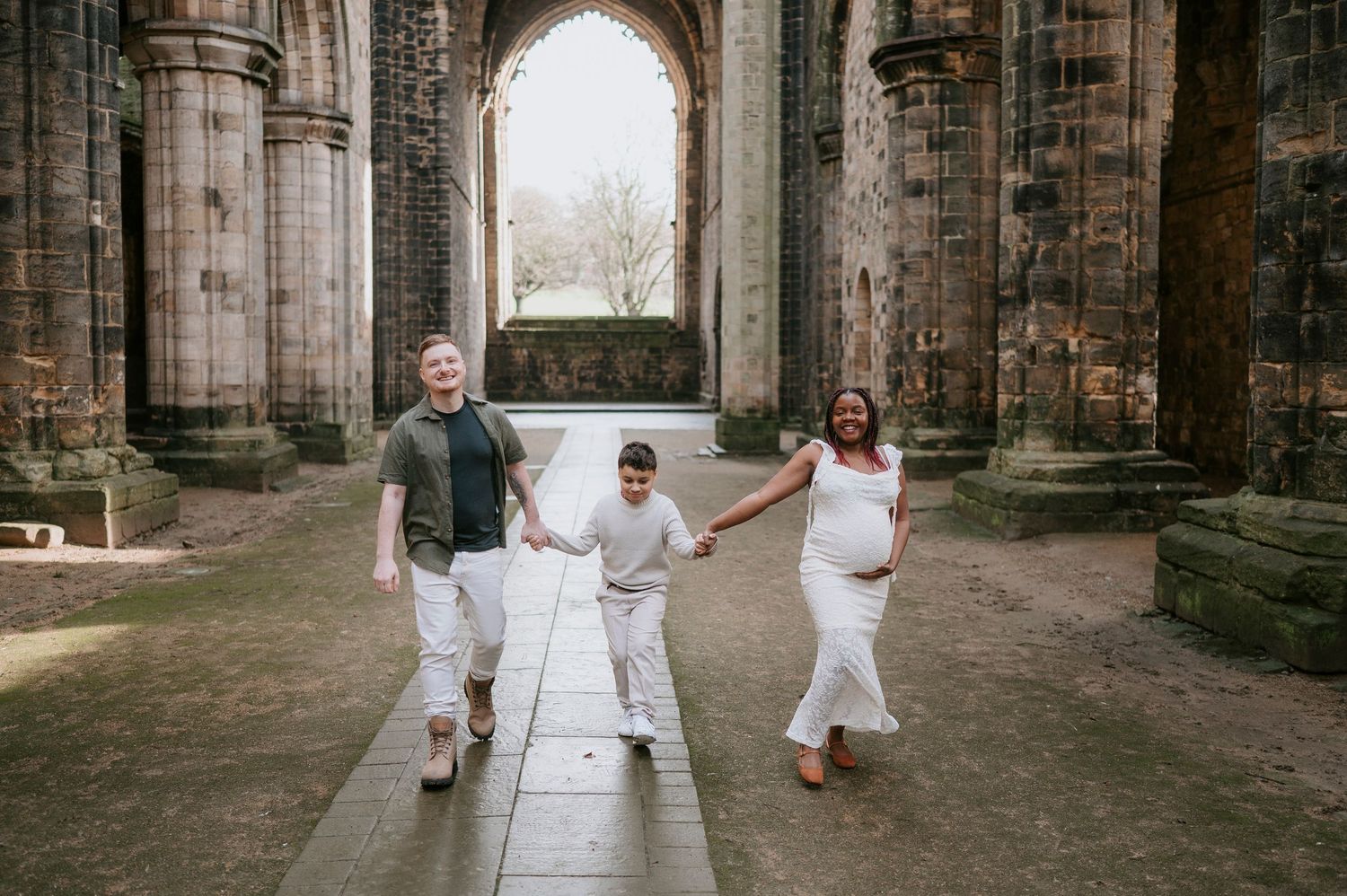 A group in white outfits holds hands while walking through the stone archways of an ancient cathedral ruin.