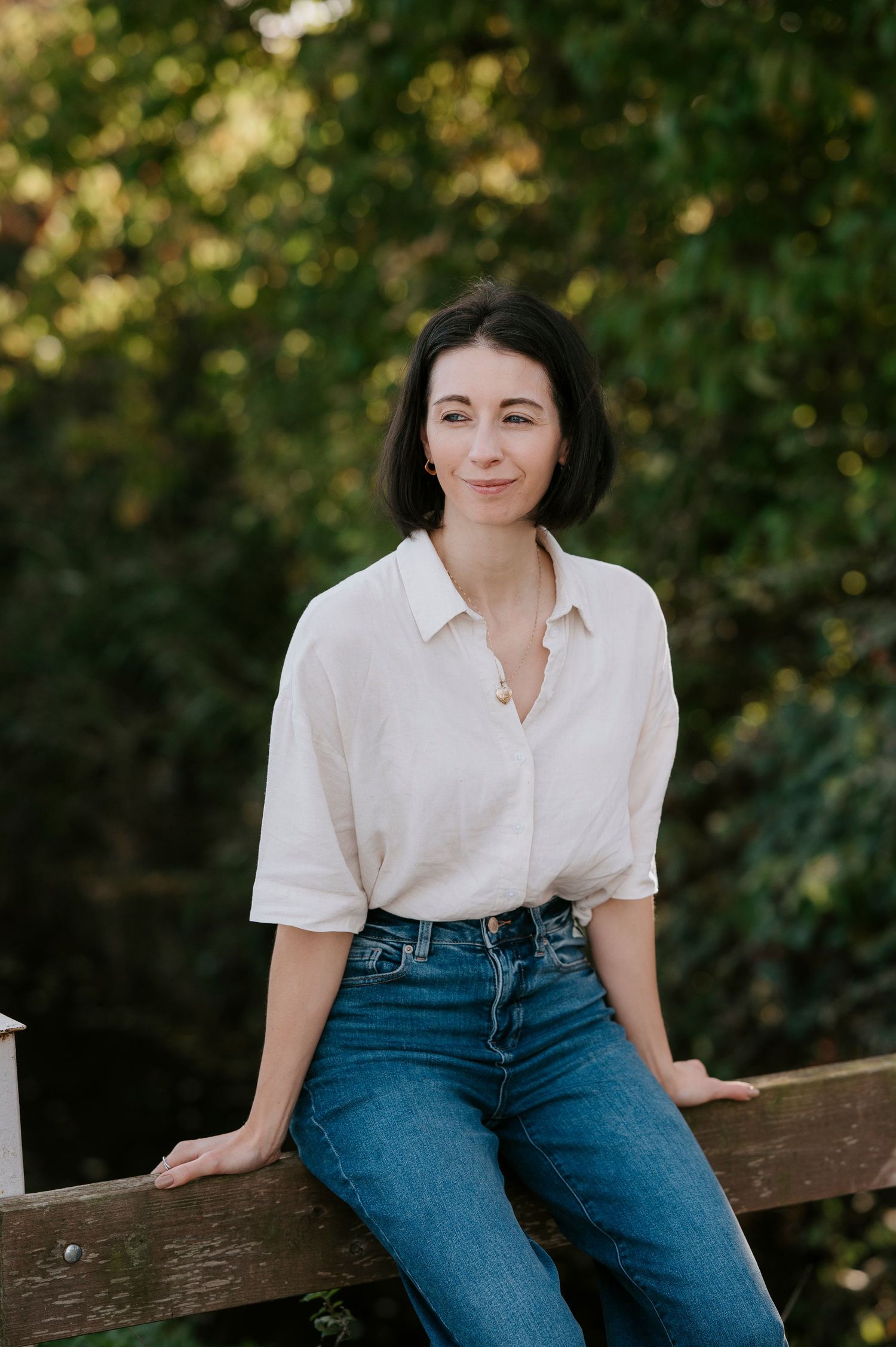 Person in casual white button-down shirt and blue jeans sits on wooden bench in outdoor setting with greenery.