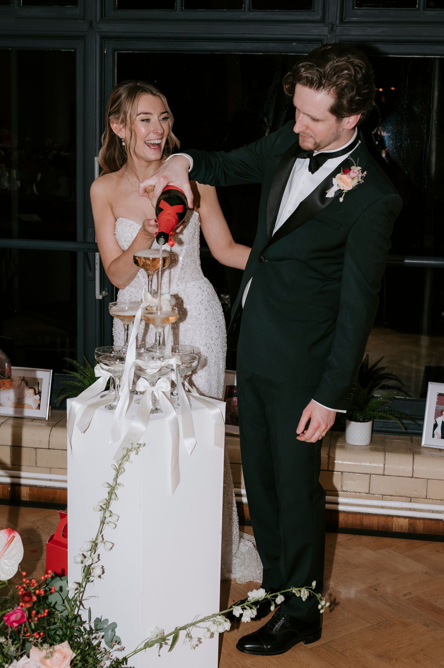 A couple shares a joyful moment during their wedding reception while pouring champagne at a decorated table.
