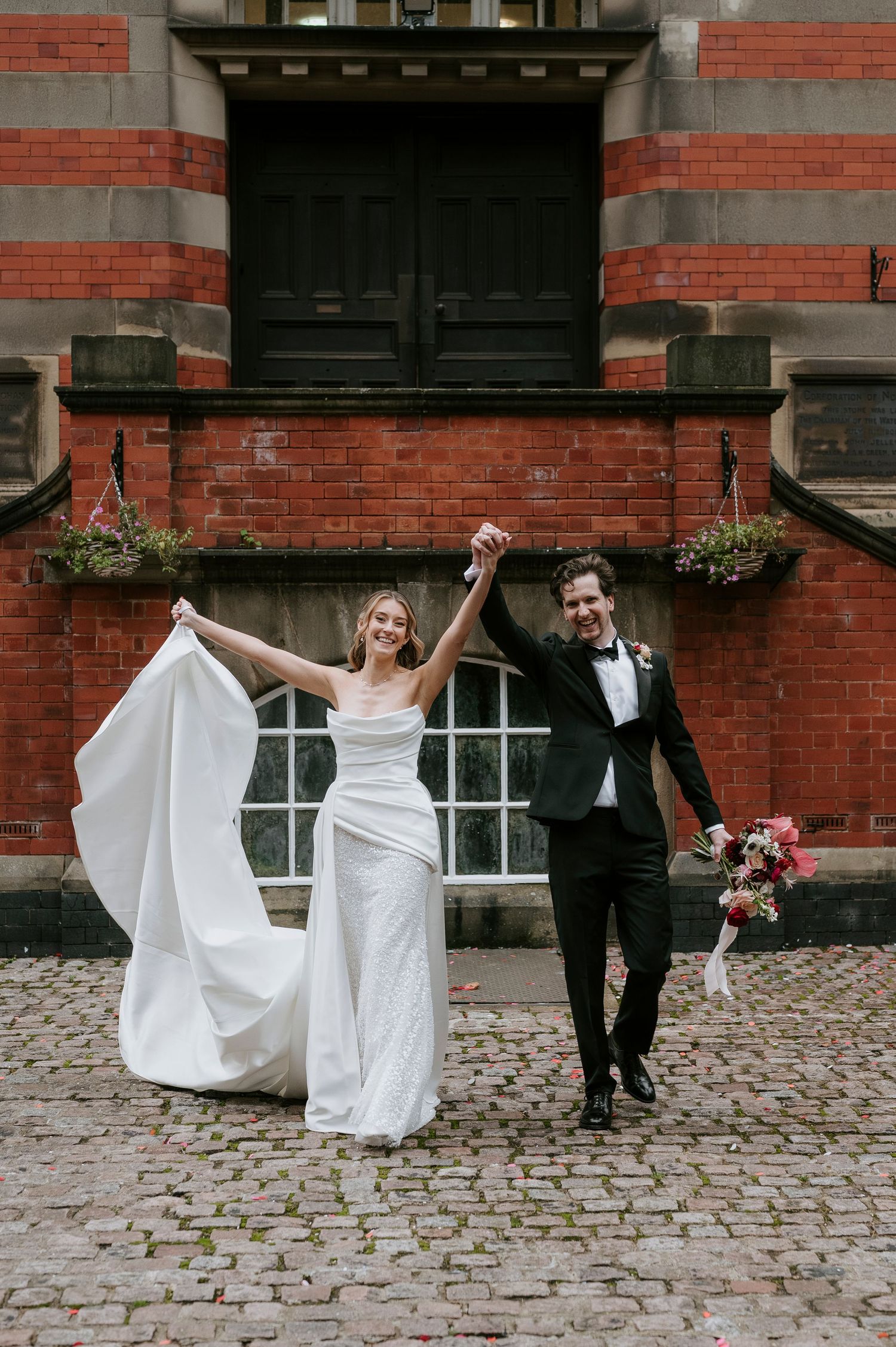 A joyful wedding couple celebrates on cobblestone street in front of historic red brick building with flowing white dress.