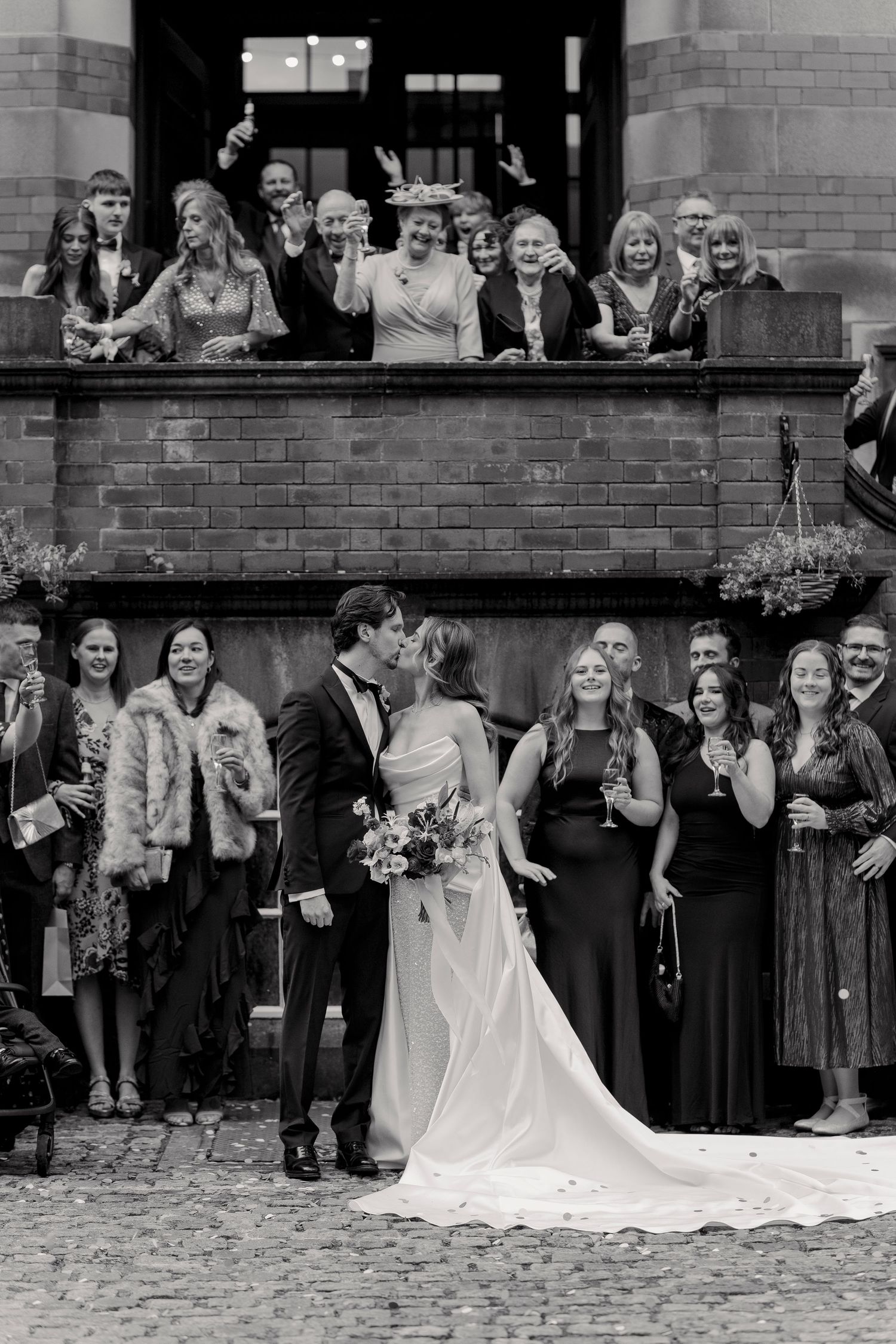 Wedding party and guests gather on multiple levels of brick building for formal group photo.