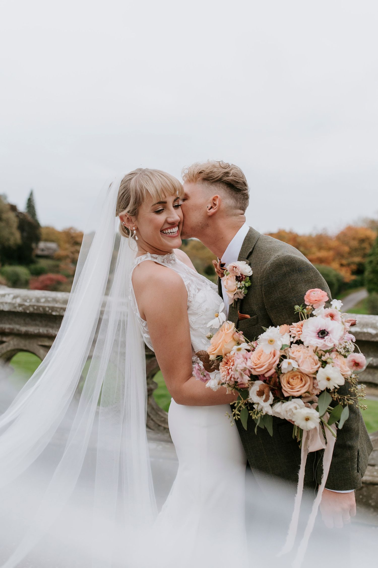 Romantic wedding couple embraces outdoors with flowing veil and pink floral bouquet in autumn setting.