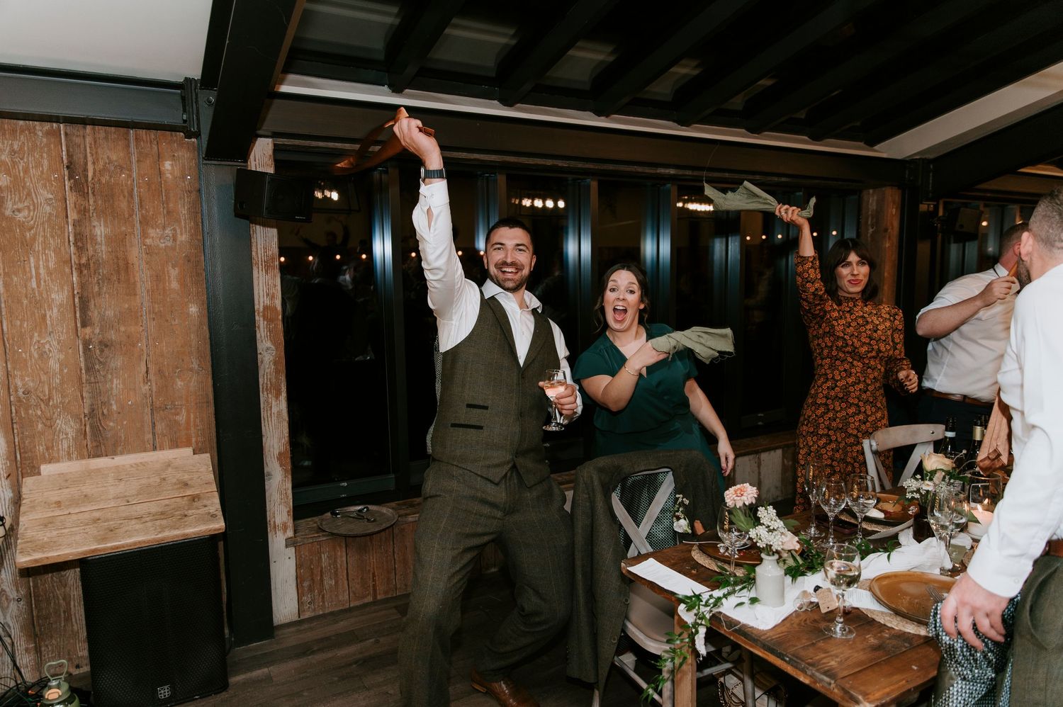 Wedding guests dance and celebrate on a rustic wooden dance floor at an indoor reception venue.