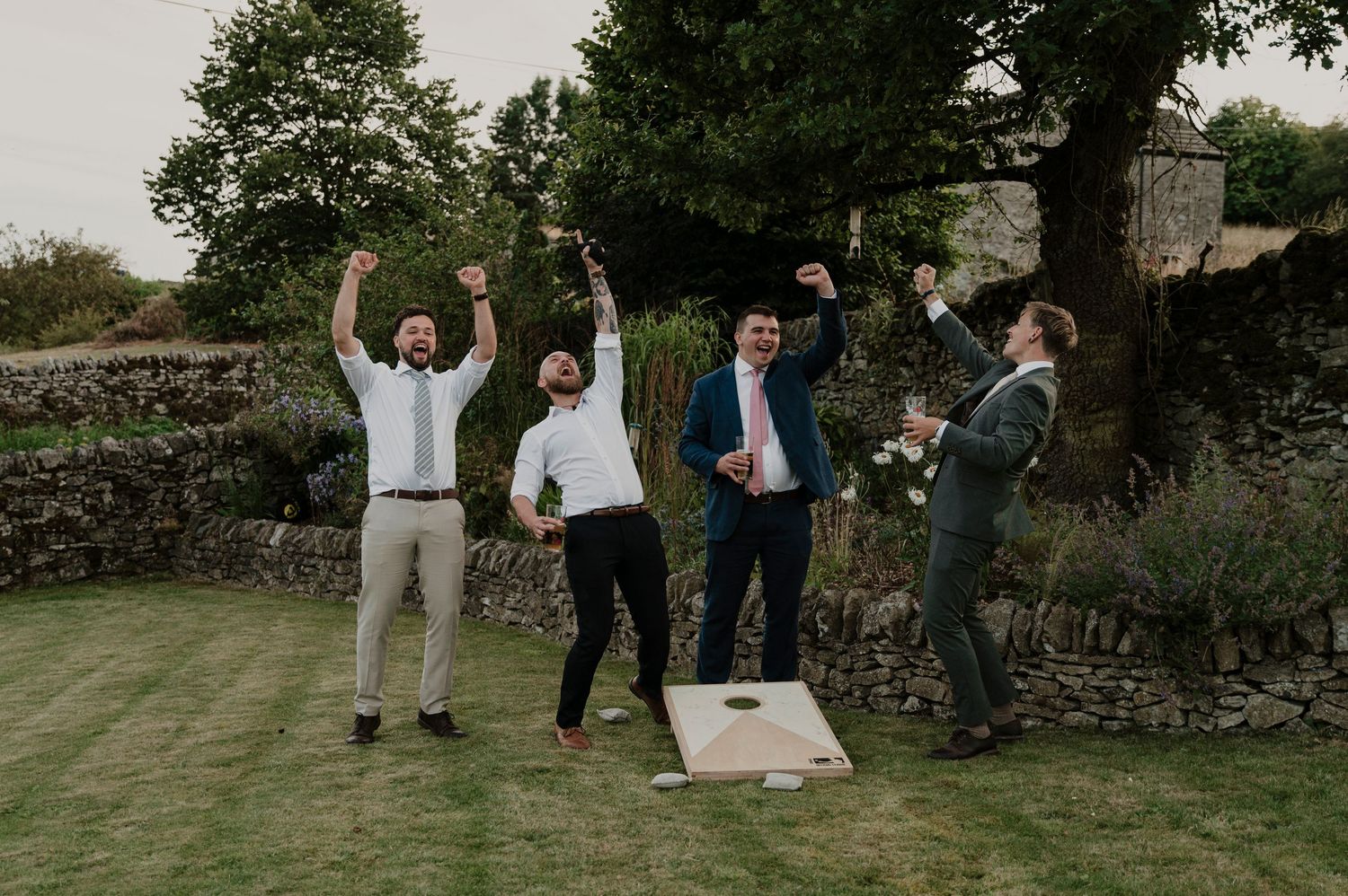 Groomsmen playing cornhole and celebrating on a garden lawn.