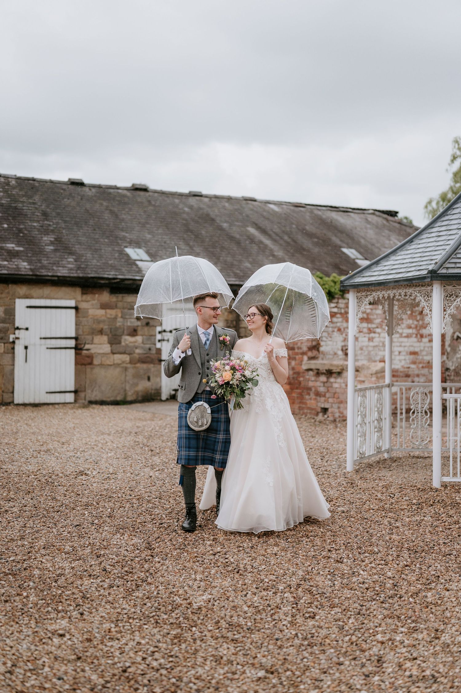 A wedding couple walks together under clear umbrellas on a gravel courtyard outside rustic stone buildings.
