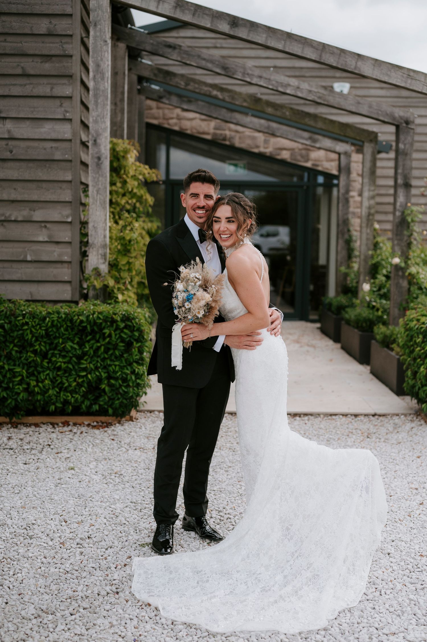 A couple poses together at their wedding on a gravel path with rustic wooden architecture and greenery in the background.