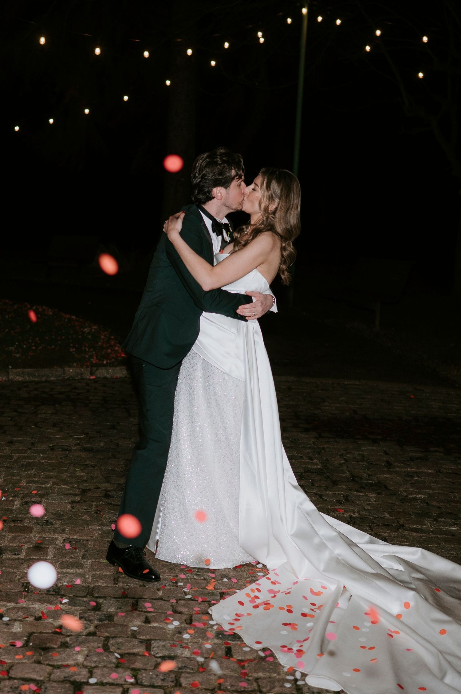 Wedding couple dancing on cobblestone patio with rose petals scattered under twinkling lights.