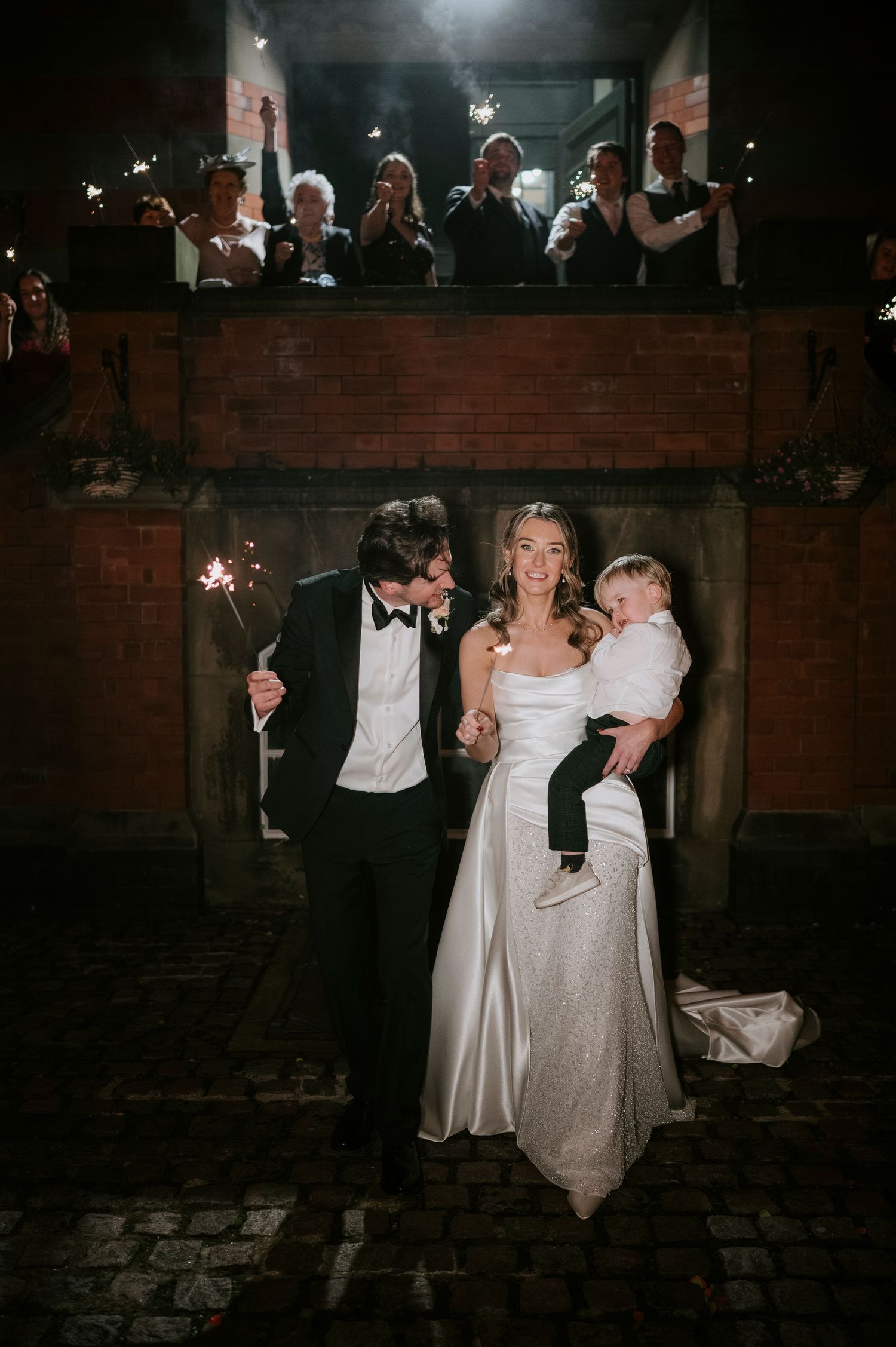 Wedding party dances together at a dimly lit evening reception with brick walls visible in the background.