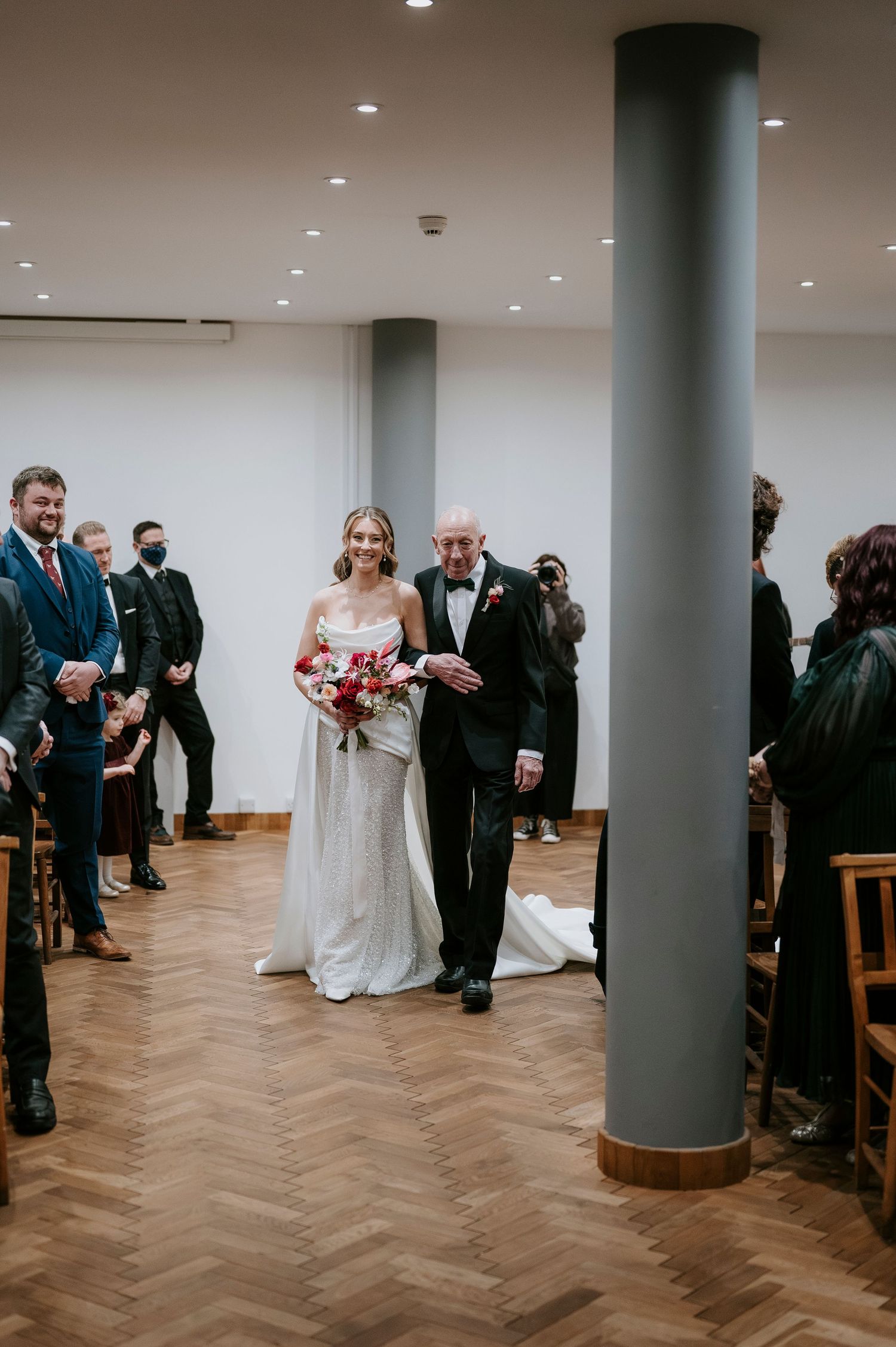 Wedding procession walking down a modern indoor venue with white columns and herringbone wood flooring.