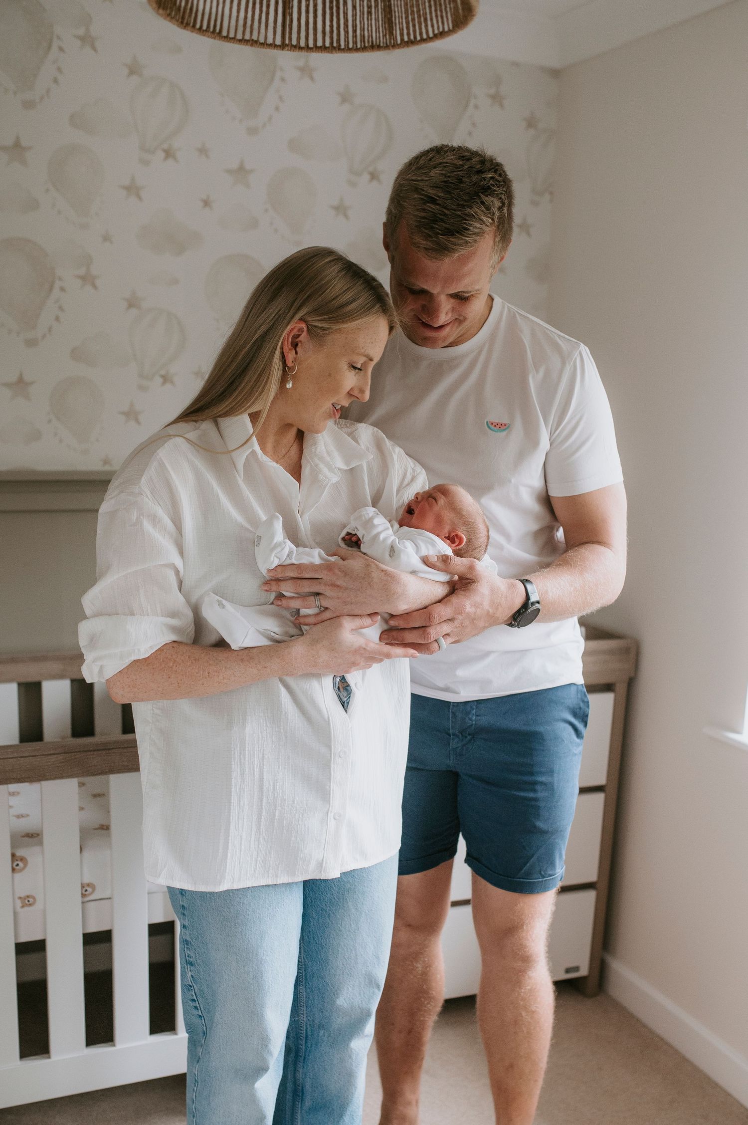 Parents in white shirts and blue bottoms hold their newborn baby in a nursery with star wallpaper.