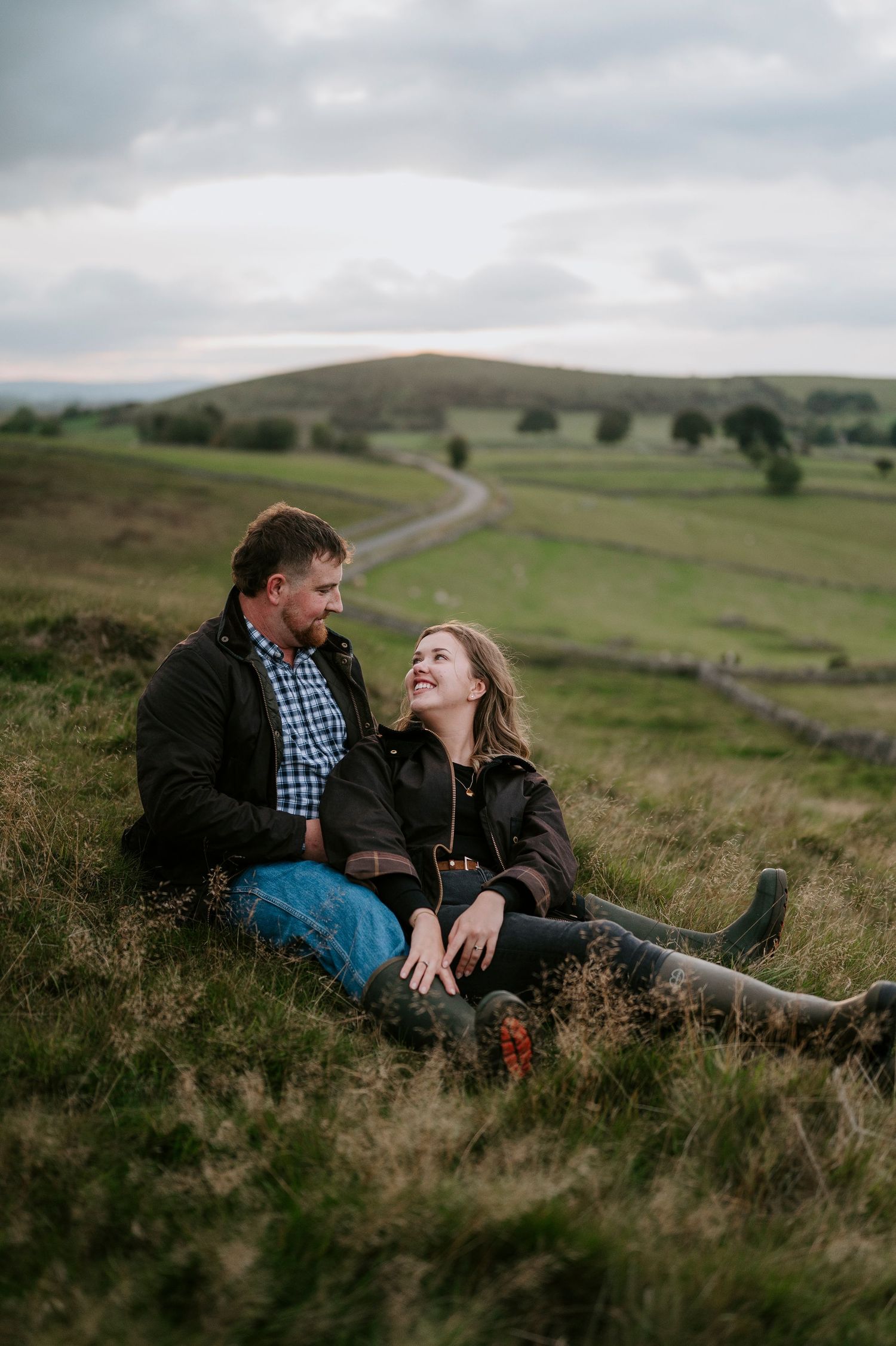 A couple shares a romantic moment while sitting on a grassy hillside overlooking rolling countryside in moody weather.