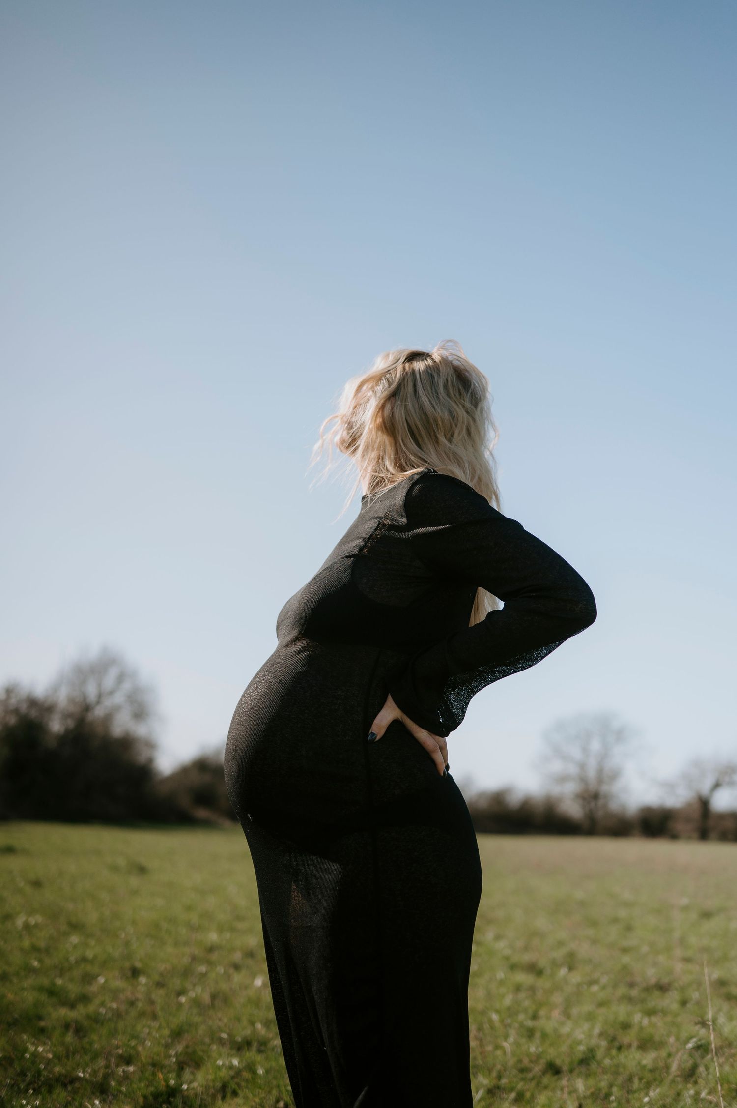 A silhouette of a pregnant figure in a black dress stands against a bright sky in an open field.