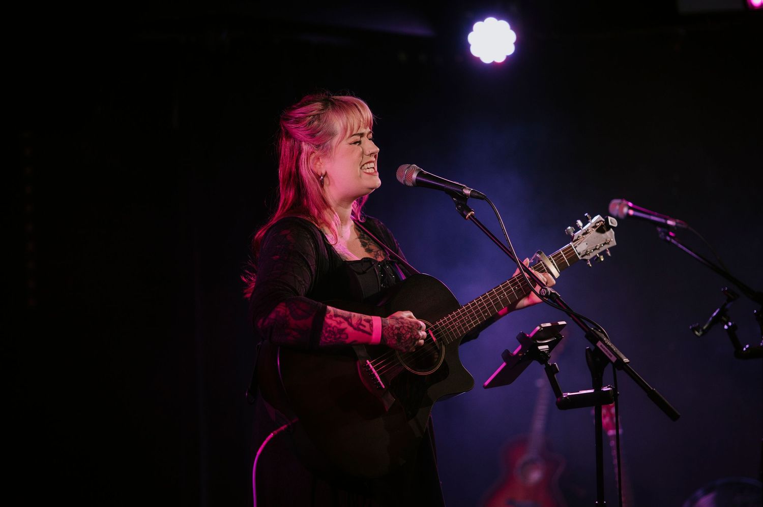 Musician performs on stage with guitar and microphone against dark background with purple lighting effects.
