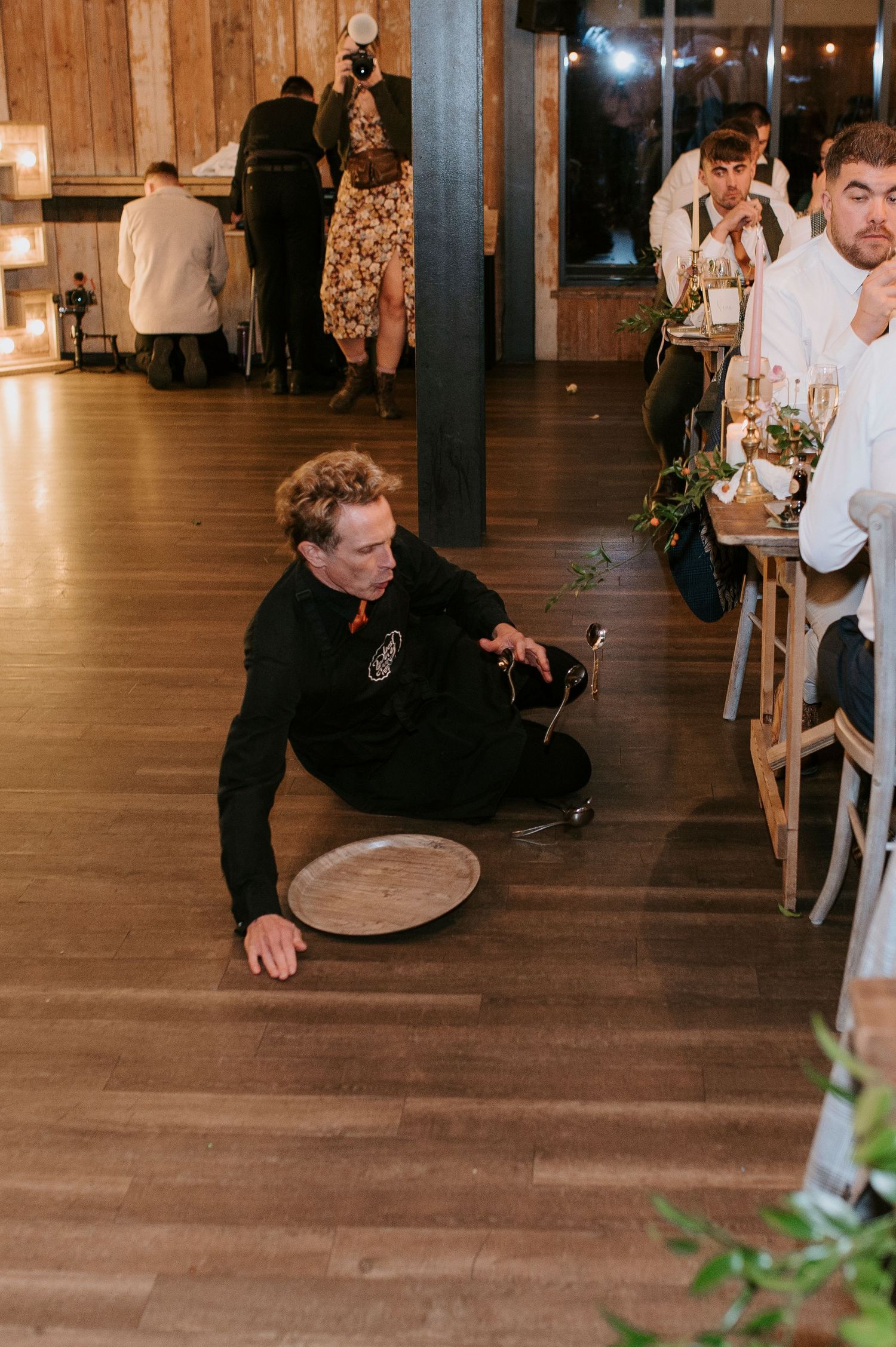 Someone in a black hoodie slips and falls while holding an empty serving tray on a wooden floor at an indoor event.