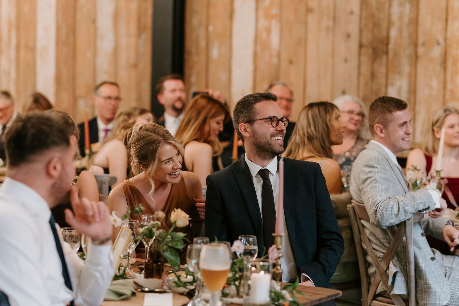 Wedding guests laugh and socialize at reception tables inside rustic wooden barn venue.